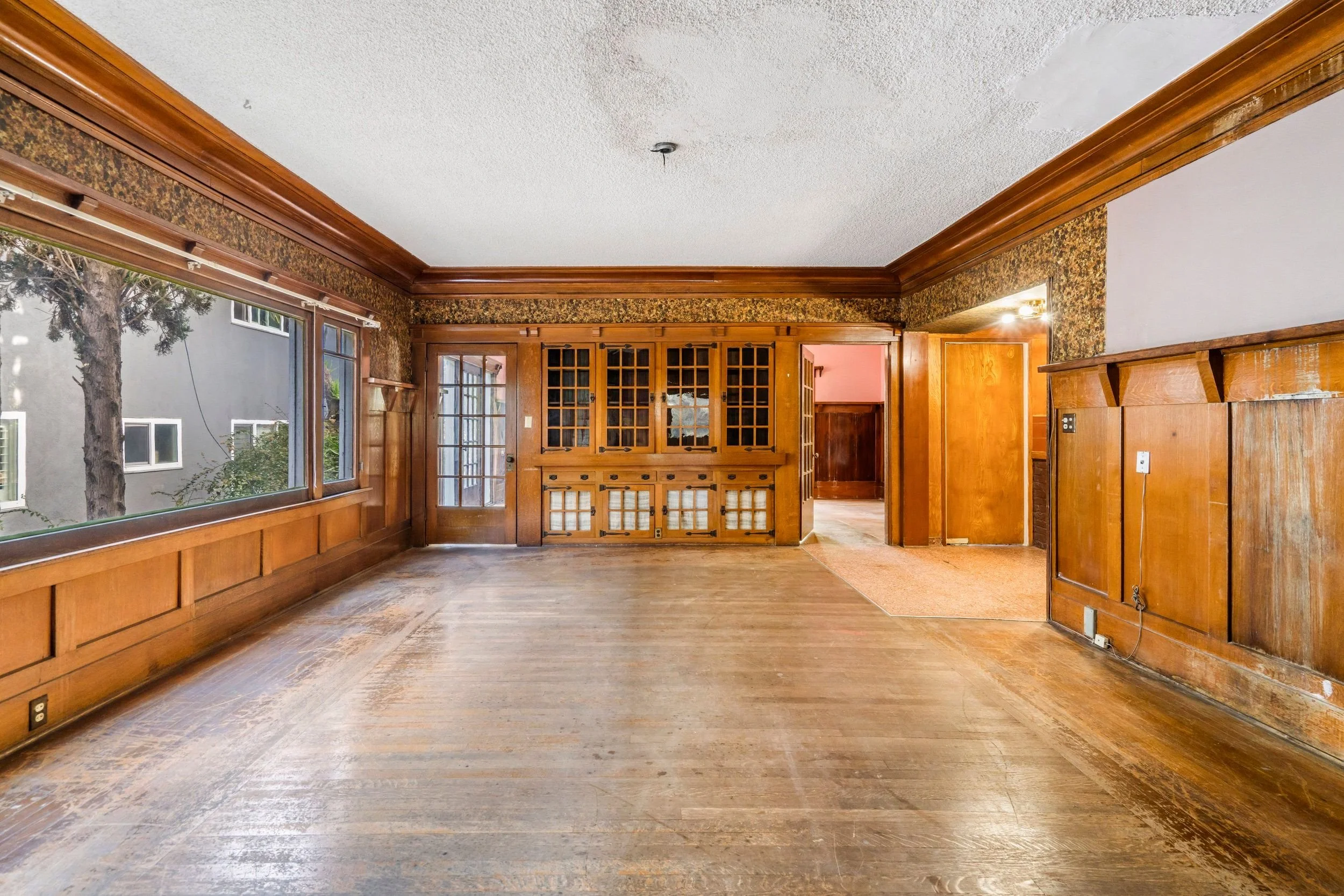 Empty living room with wooden flooring, wood-paneled walls, large windows, and a built-in bookshelf.