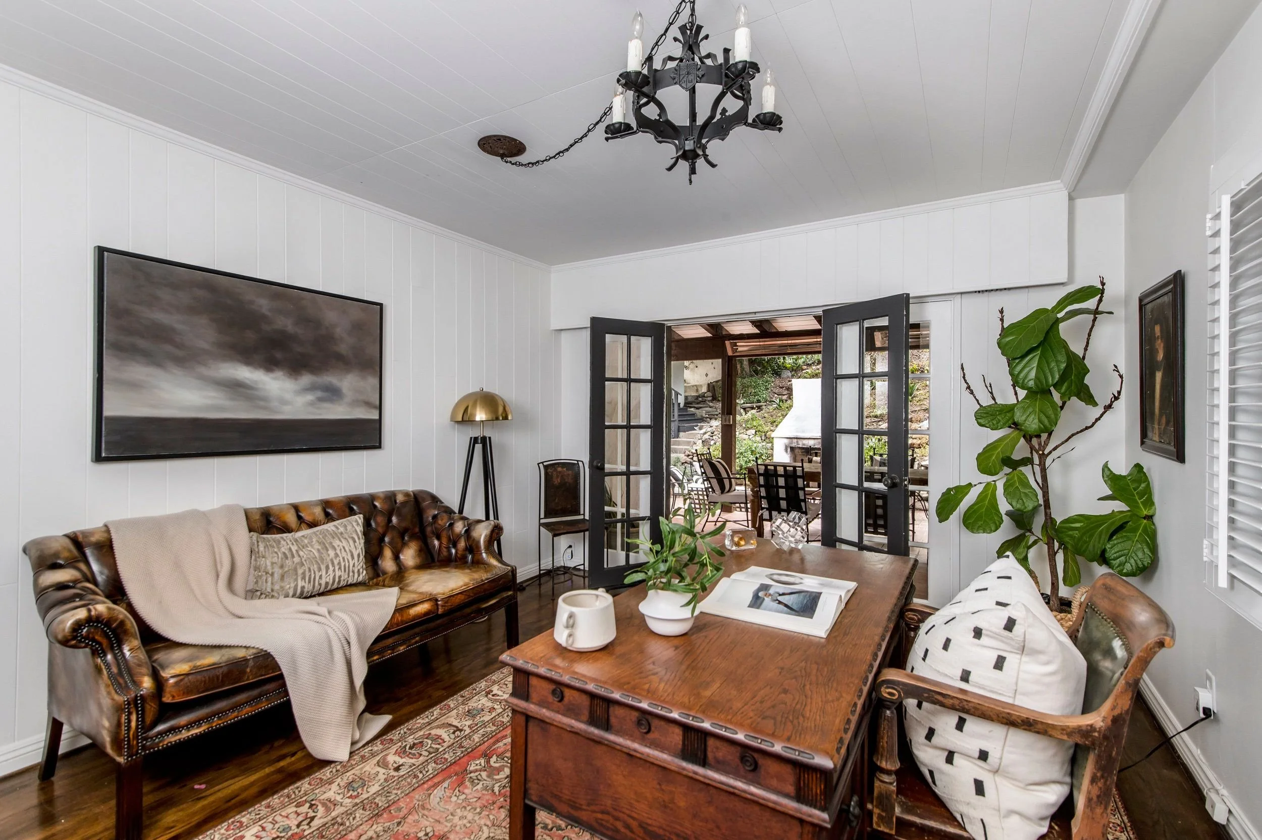 Living room with brown leather sofa, beige throw blanket and patterned pillows, wooden coffee table with books and plants, black chandelier hanging from white ceiling, white paneled walls, large window and door leading to outdoor patio with dining ta