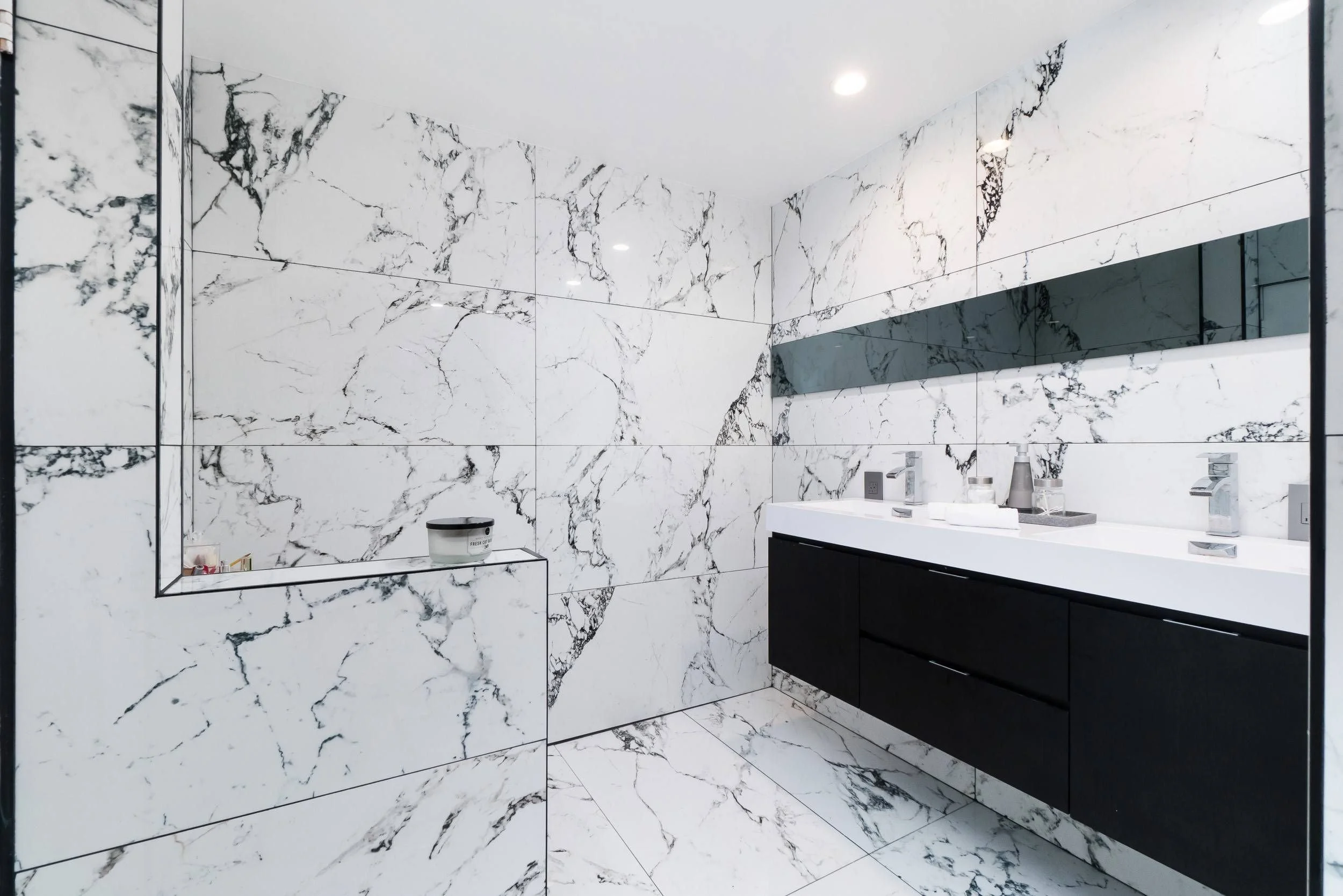 Modern bathroom with white marble walls and floor featuring black veining, a large mirror, a white double sink vanity with black base drawers, and various toiletries on the sink counter.