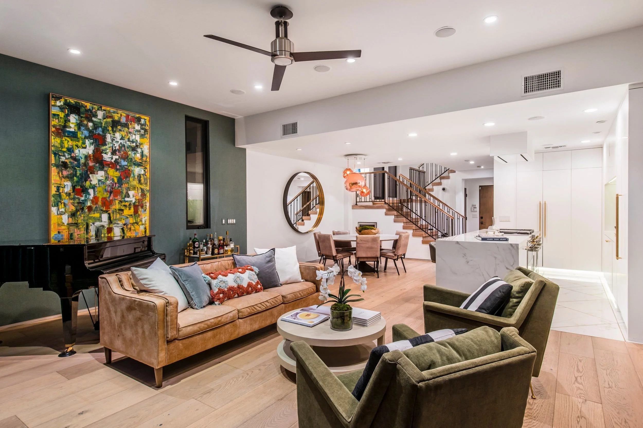 Modern open-concept living room and dining area with contemporary decor, featuring a leather sofa, green armchair, abstract wall art, a round mirror, and a staircase with black metal railing.