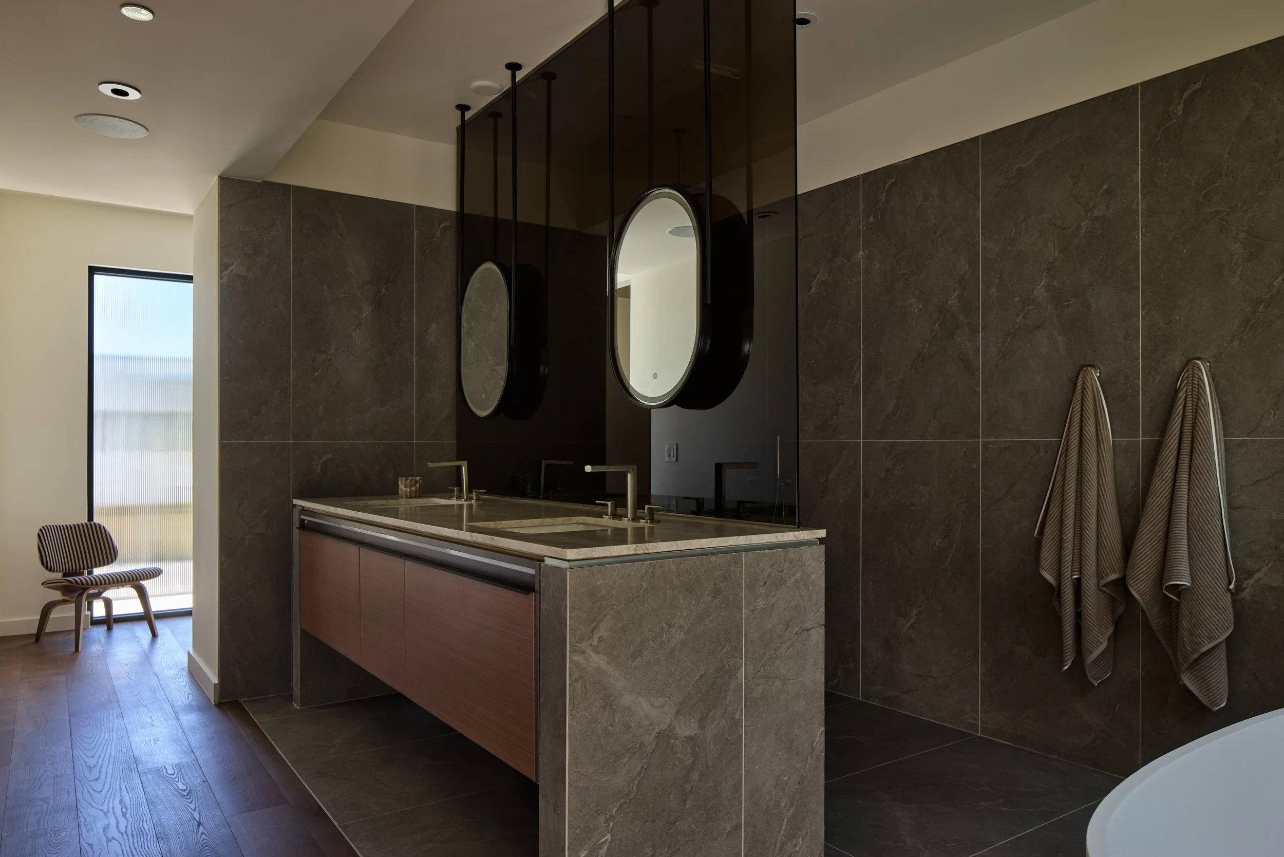 Modern bathroom with dark marble tiles, a double vanity with a marble countertop, two oval mirrors with black frames, and two hand towels hanging on the wall. A chair with striped fabric and a window are also visible.