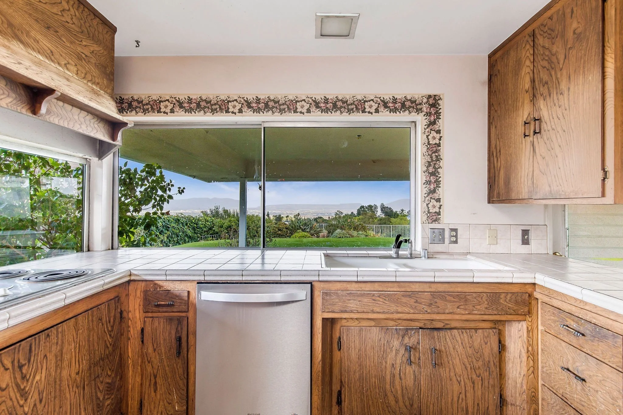 View of a kitchen with wooden cabinets, tiled white countertops, a large window showing greenery and distant mountains, and a ceiling with a skylight.
