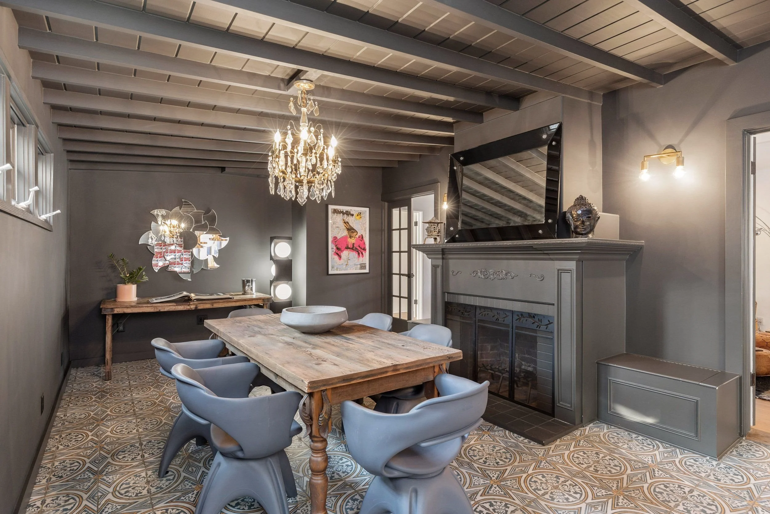 A cozy dining room with a wooden table surrounded by eight modern gray chairs. The room features patterned tile flooring, a gray-painted wall, a decorative mirror, a chandelier, and various wall art. A fireplace with a gray mantel and decorative scul