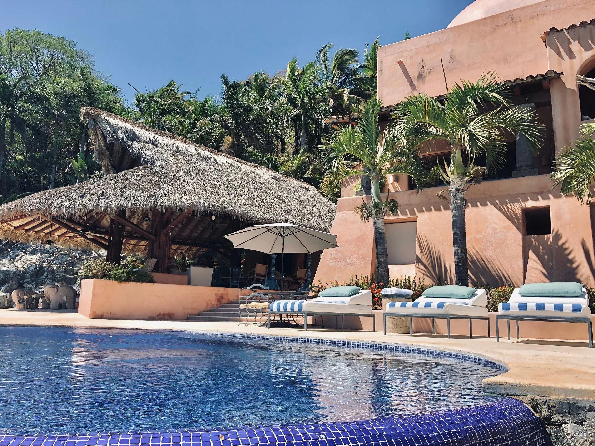 A swimming pool area at a resort with lounge chairs, umbrellas, and palm trees, with a pink stucco building and thatched roof hut in the background under a blue sky.