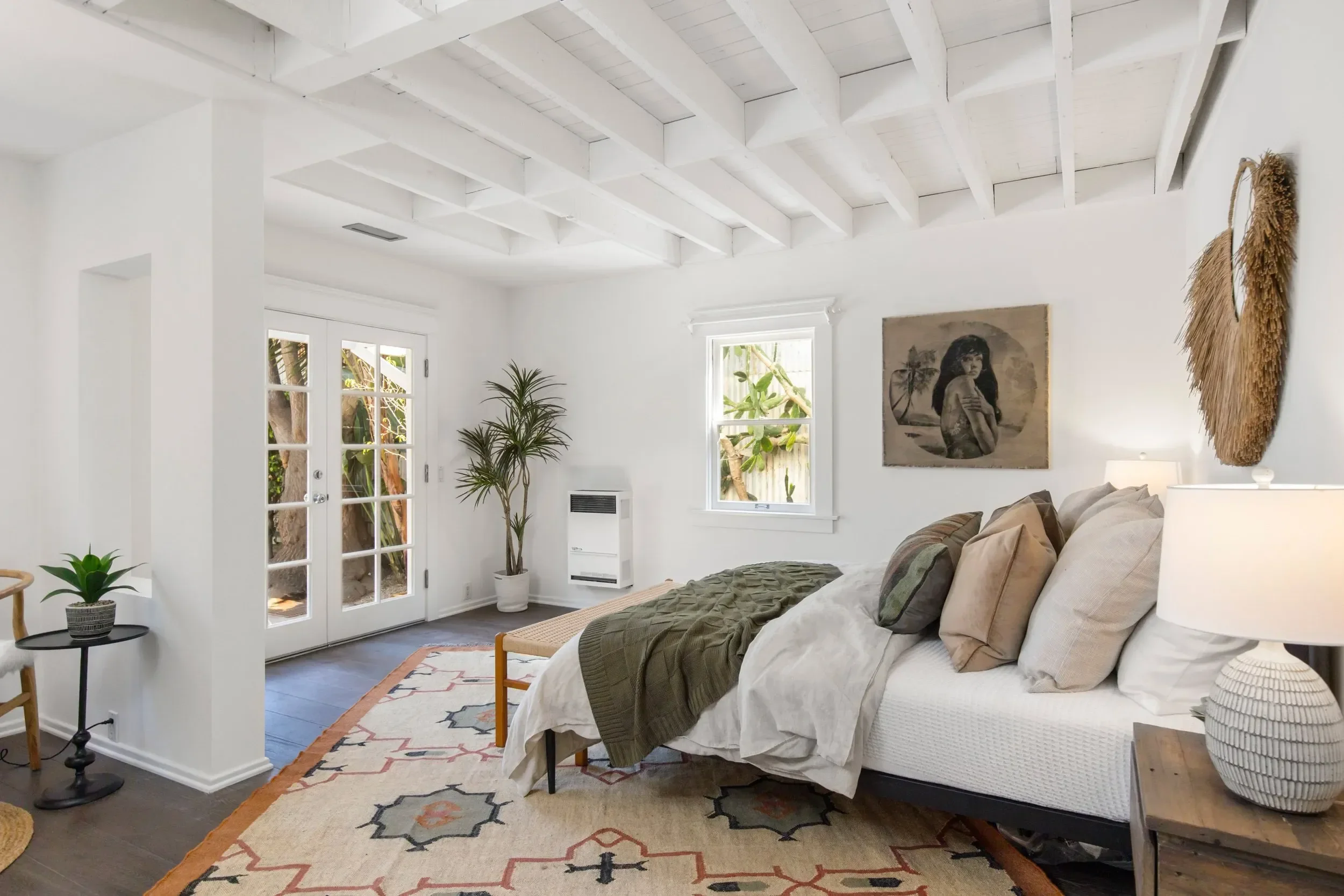 A bright living room with white walls, a white ceiling with exposed beams, and large windows. There is a beige sofa with pillows, a wooden bench at the foot of the sofa, and various houseplants. The room features wall art, a small table with a plant,