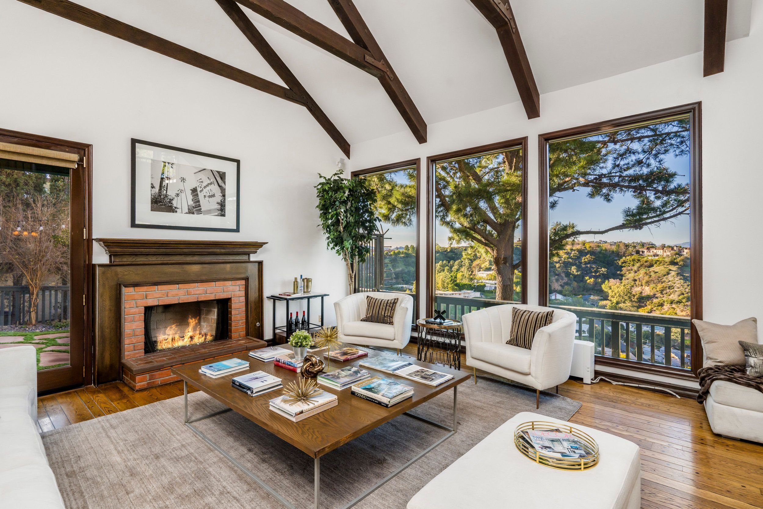 Living room with a fireplace, white sofas, a large wooden coffee table with magazines, tall windows with scenic views of trees, and a potted plant.