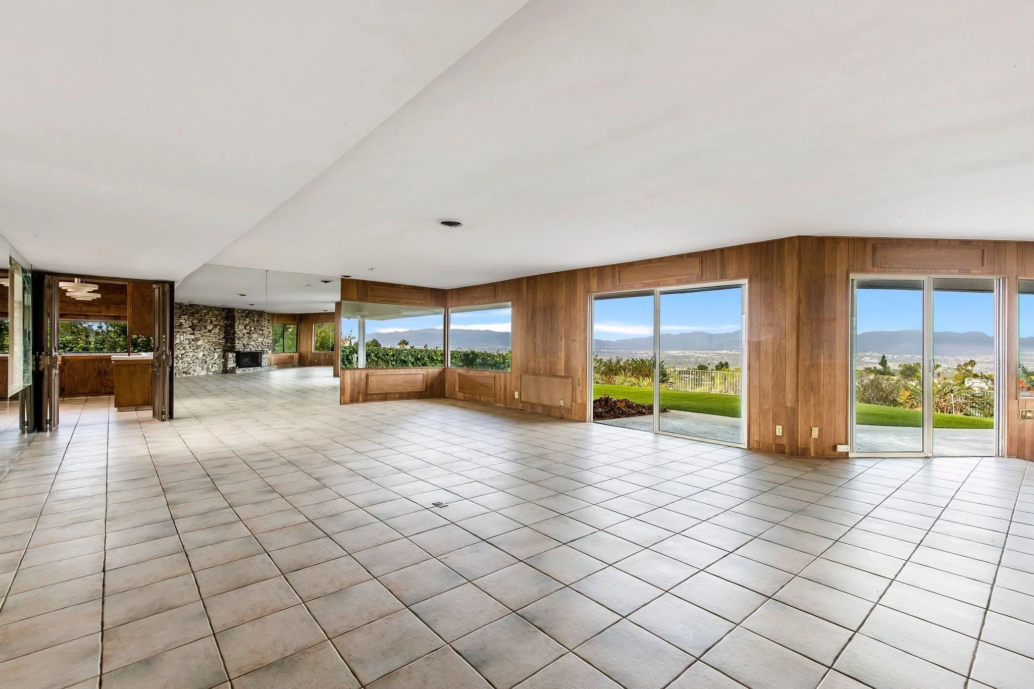 Empty living room with large windows and sliding glass doors overlooking a garden and mountains, wood-paneled walls, tiled floor, and a ceiling with a smoke detector.