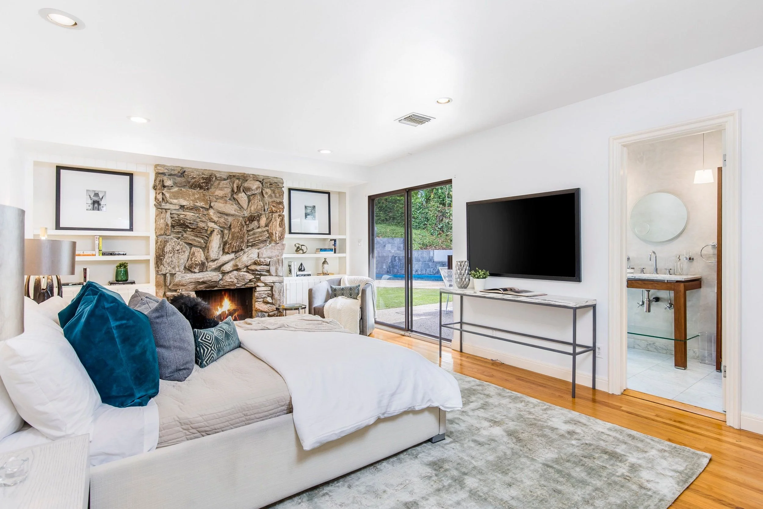 A living room with a white sofa adorned with blue and gray pillows, a stone fireplace with a fire, a black framed photograph above the fireplace, white built-in shelves on each side with books and decorative items, a large flat-screen TV mounted on t