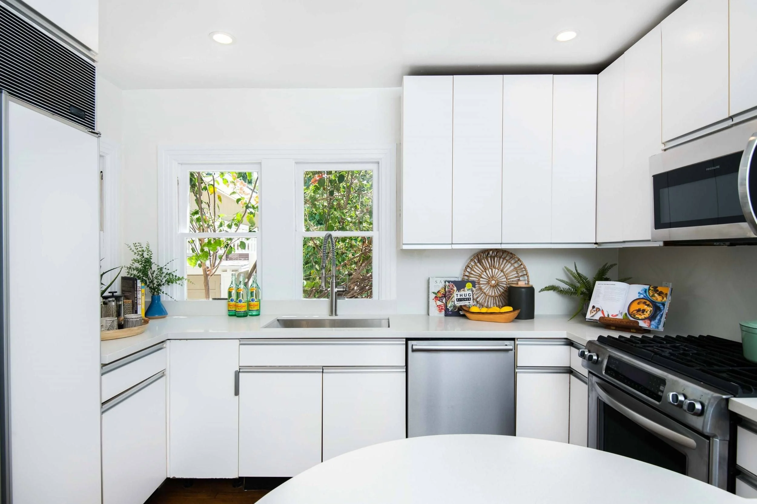 Modern white kitchen with two windows above the sink, greenery visible outside, and various kitchen items like bottles, books, and a fruit bowl on the counter.