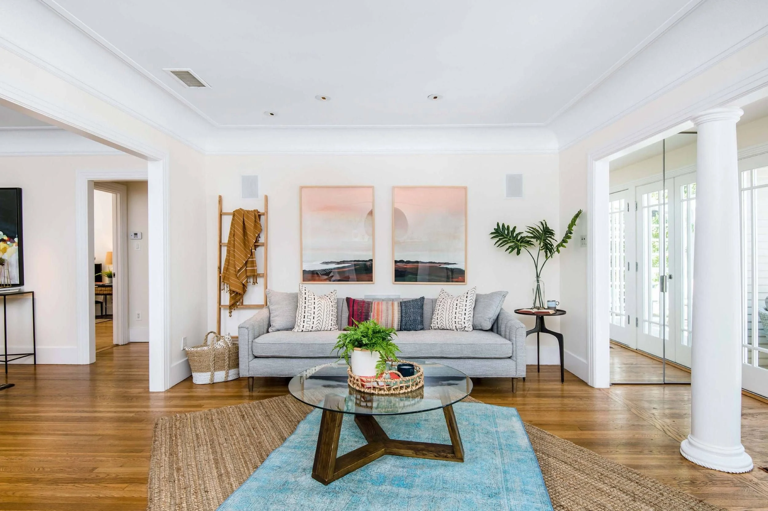 Living room with gray sofa, wooden floor, glass coffee table, blue and beige rugs, wall art, and large windows.