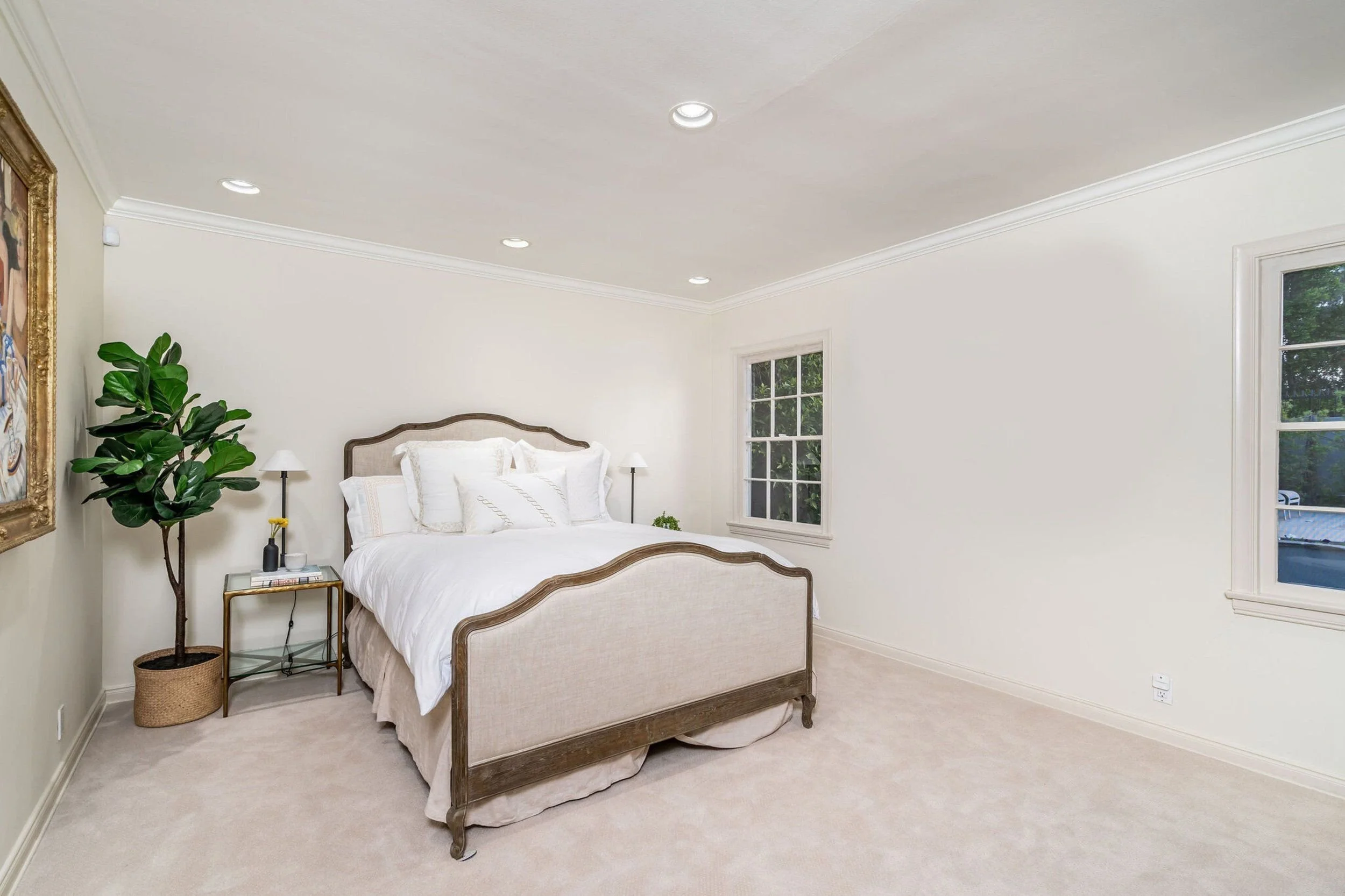 A cozy bedroom with a large bed with a beige upholstered headboard and white bedding, flanked by two small white bedside tables with lamps. A large green potted plant is placed in a wicker basket near the corner, and a portrait hangs on the wall to t