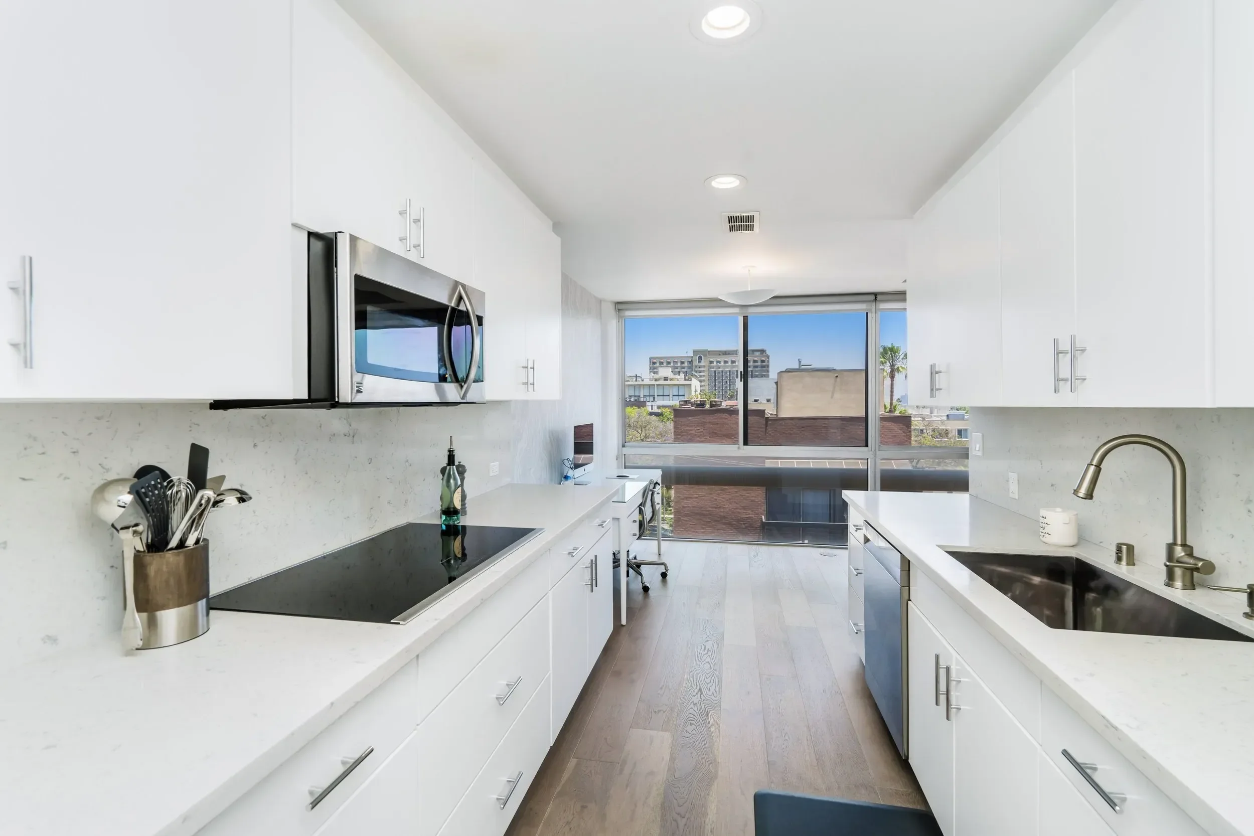 Modern white kitchen with large window showing city buildings, with countertops, cabinets, and stainless steel appliances.