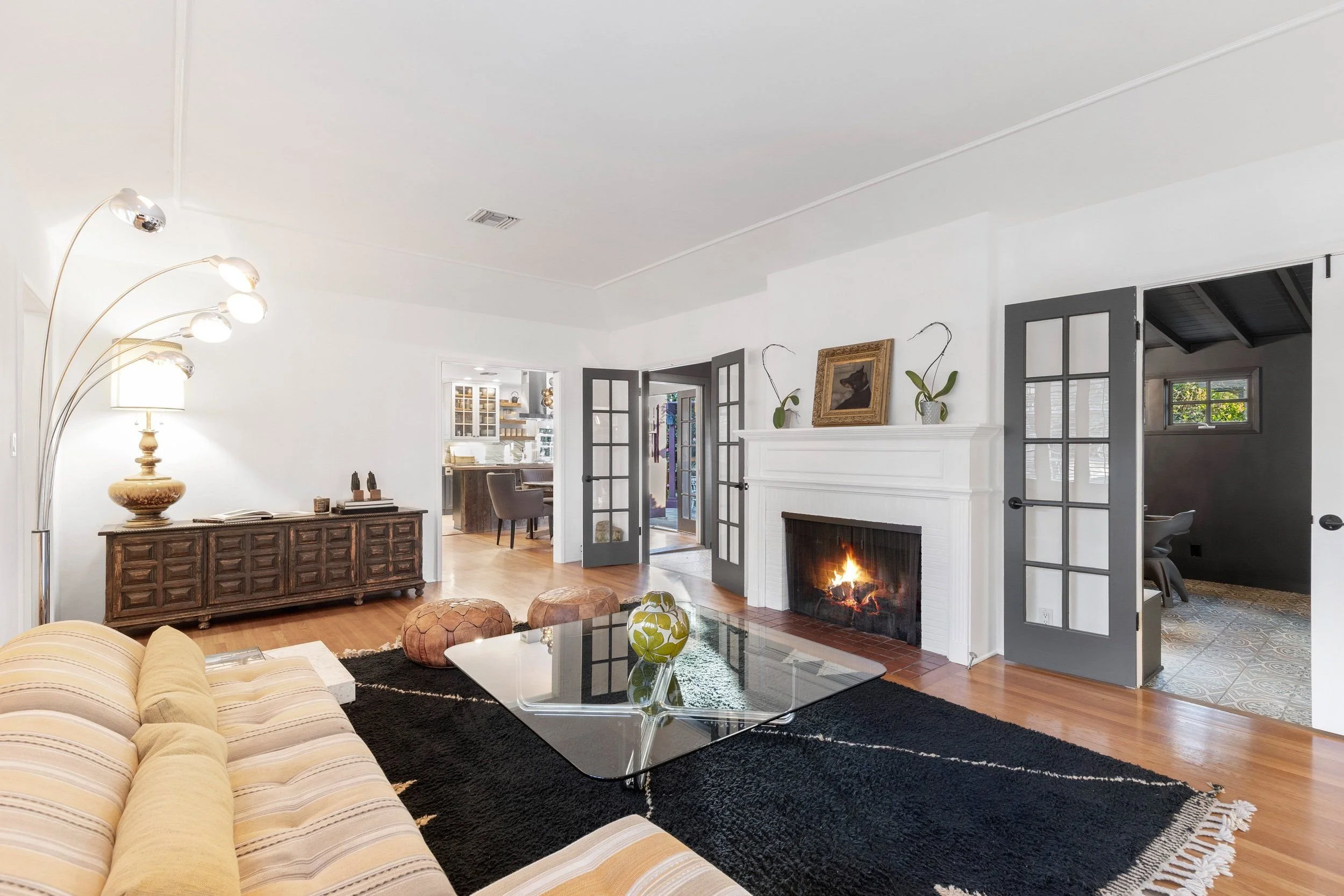 Living room with beige striped sofa, black rug, glass coffee table, wooden sideboard, and fireplace with fire, decorated with framed art and potted plants.