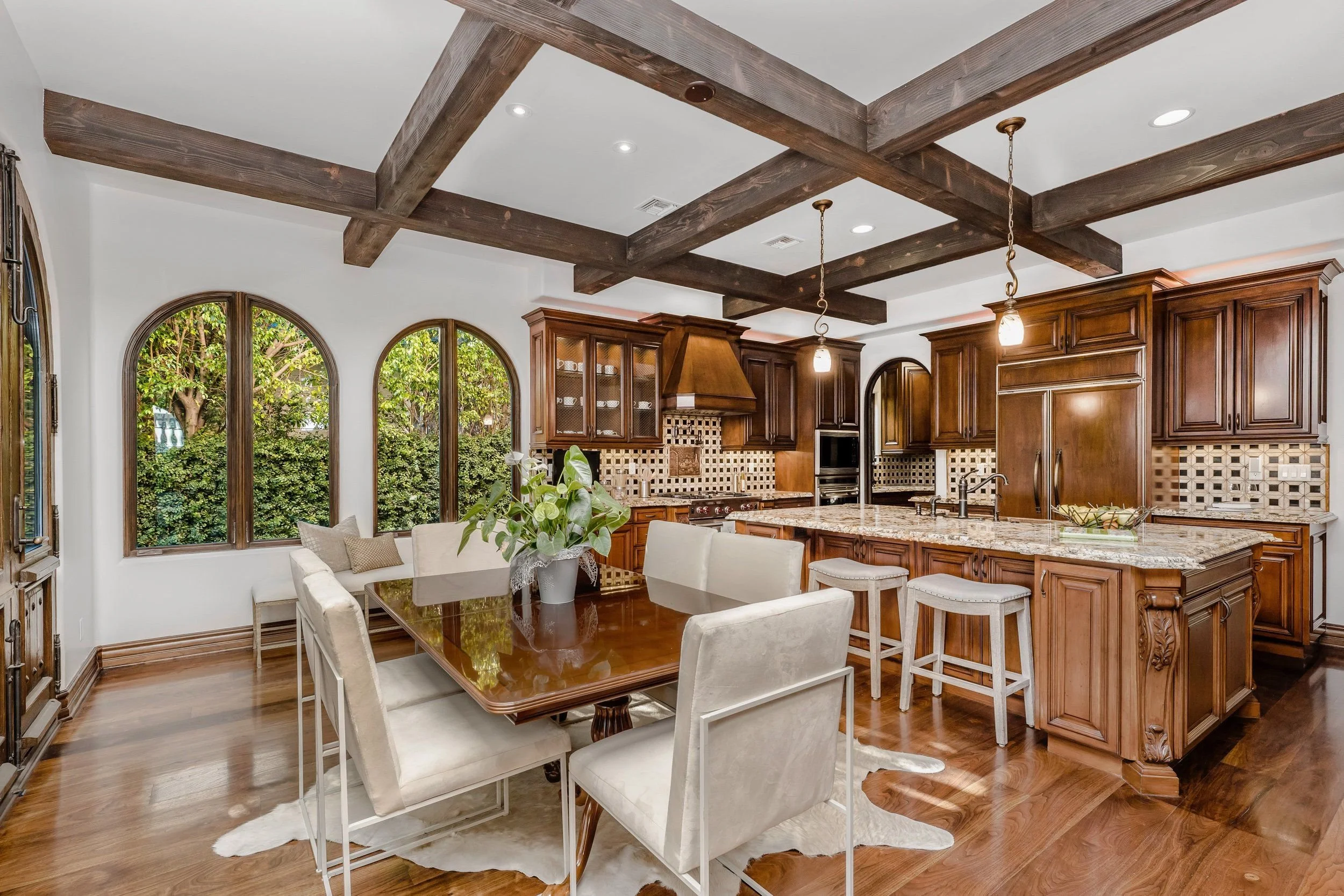 Modern kitchen with dark wood cabinets, granite countertops, and a decorative tiled backsplash. There are large arched windows overlooking green trees. A dining table with white chairs is in the foreground, and exposed wooden beams are on the ceiling