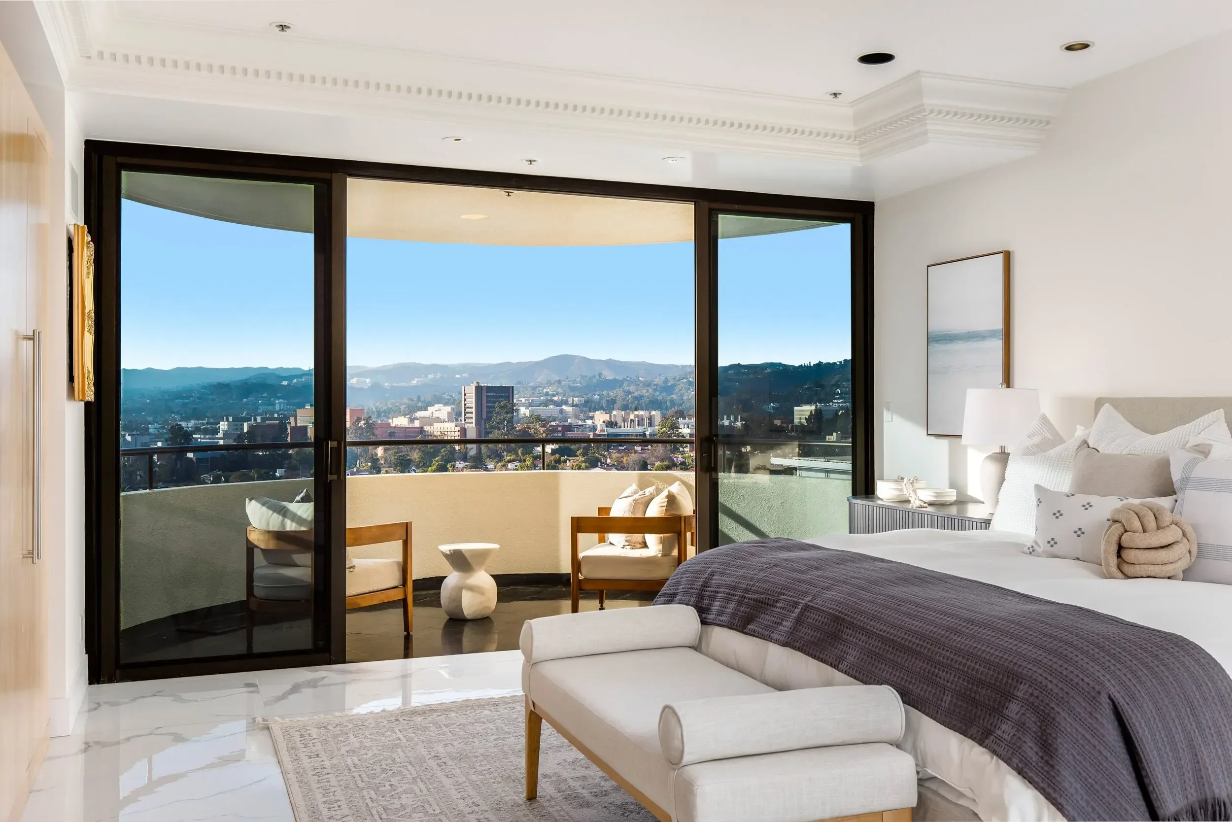 A bedroom with large glass sliding doors leading to a balcony with a city and mountain view, white walls, bed with gray bedding and white pillows, a nightstand with a lamp, and modern decor.