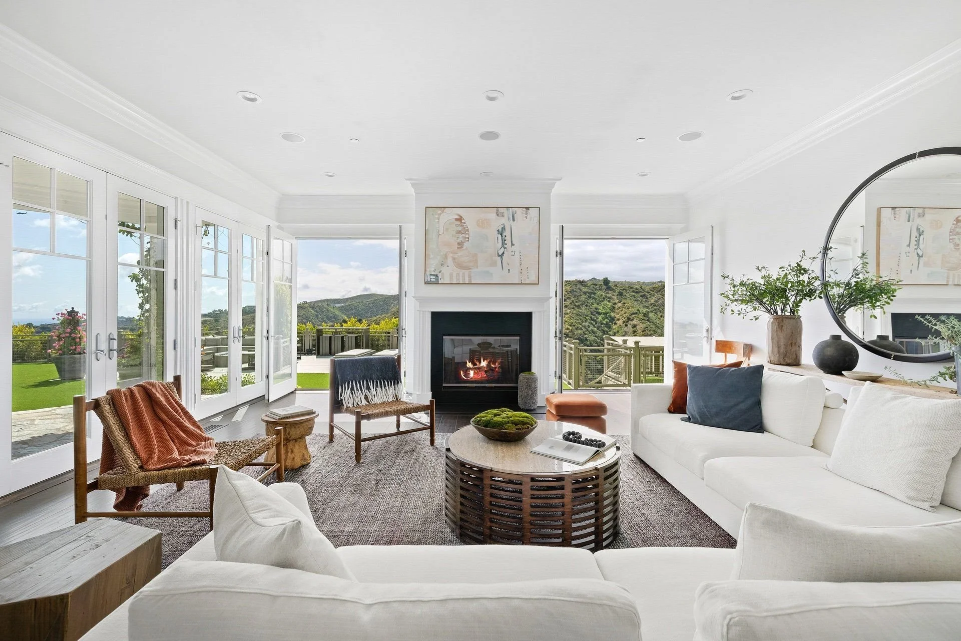 Bright living room with large windows and view of green hills, white walls, fireplace, modern art, round mirror, green plants, white sofa with pillows, wooden chairs, coffee table, and outdoor balcony.