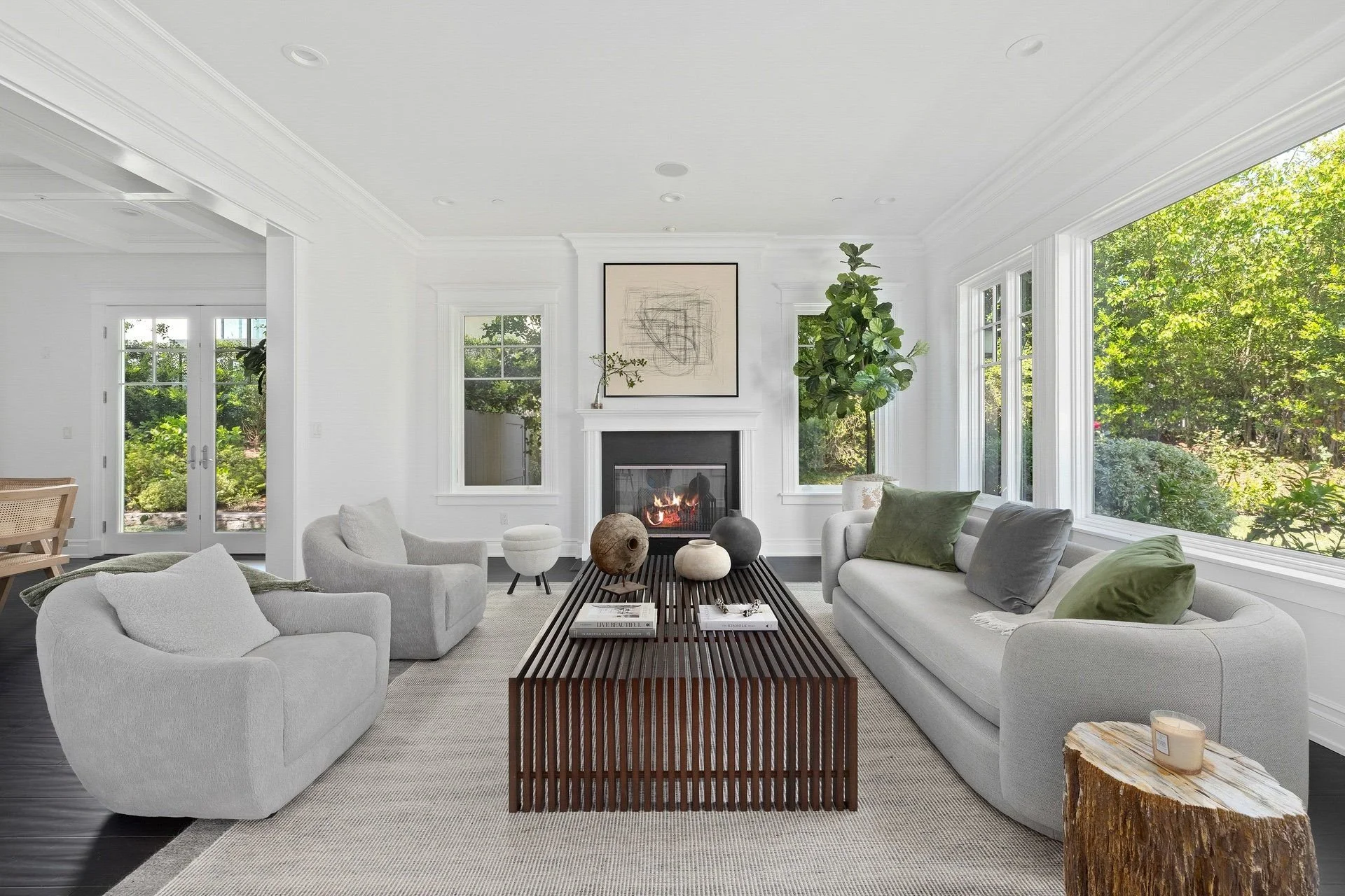 Bright living room with white walls, large windows, and a fireplace. Contains various modern furniture, including a white sofa with green and gray pillows, a curved armchair, and a wooden coffee table with decorative vases. There is a fireplace with 