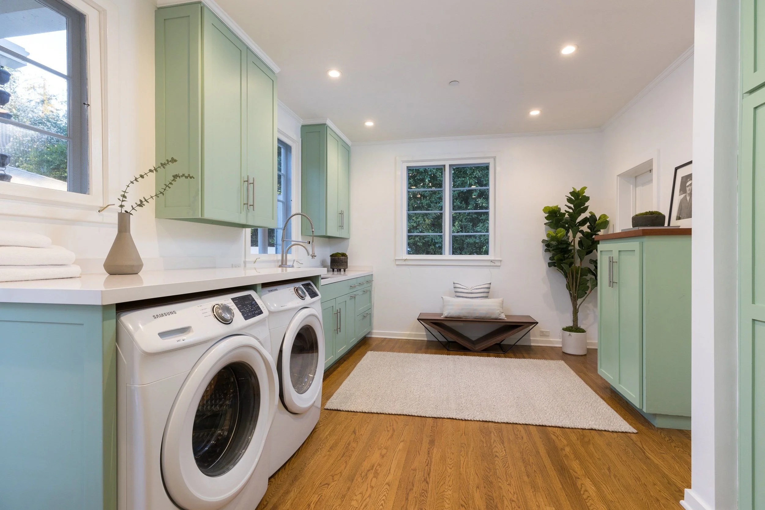 Laundry room with mint green cabinets, white washer and dryer, wood flooring, and a large window showing trees outside.