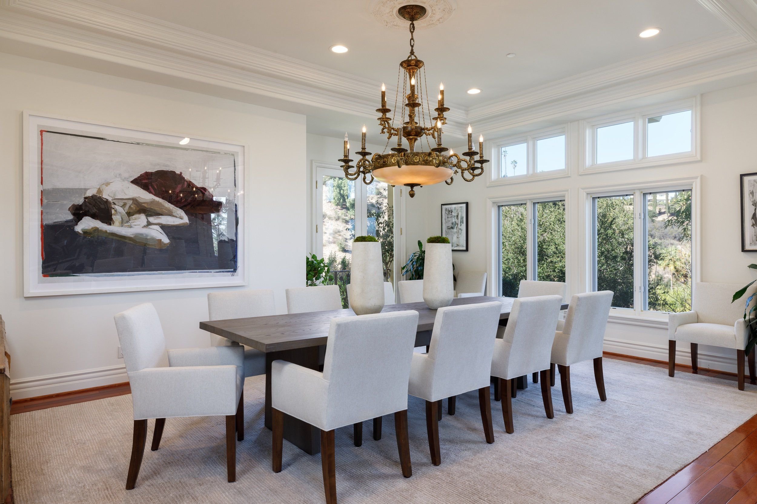 Dining room with a large wooden table, white upholstered chairs, a chandelier, and multiple windows showing greenery outside.