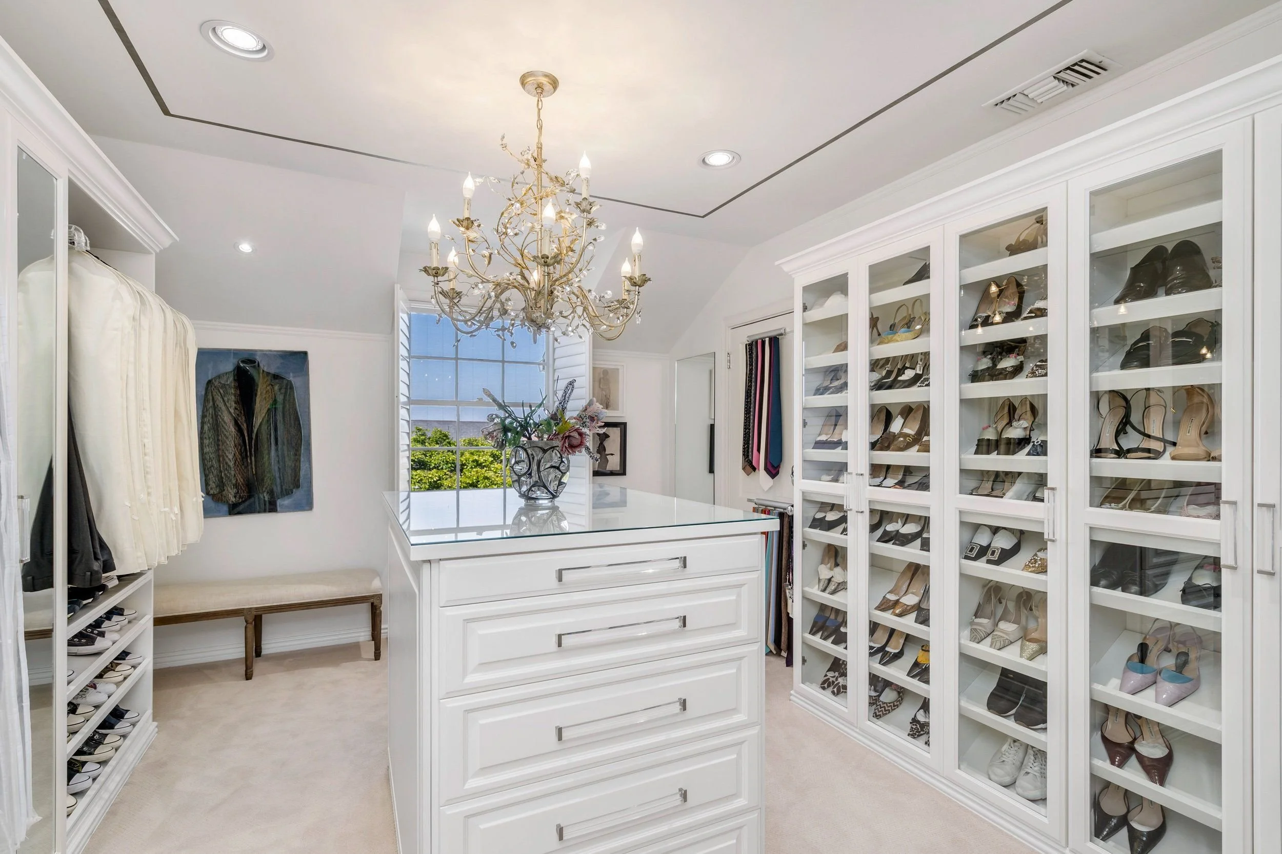 A walk-in closet with white cabinetry, glass-front shoe display shelves filled with various shoes, a white dresser with drawers, a window with shutters, a chandelier hanging from the ceiling, and a few scattered clothing items and picture frames.