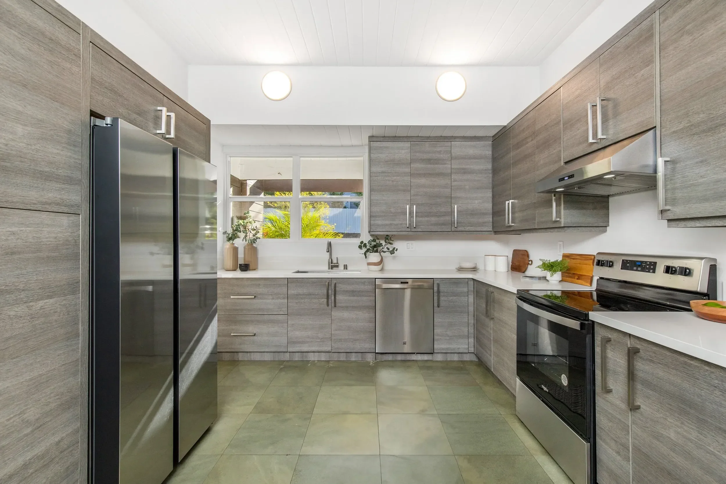 Modern kitchen with gray wood cabinets, stainless steel appliances, and a window with outdoor greenery.
