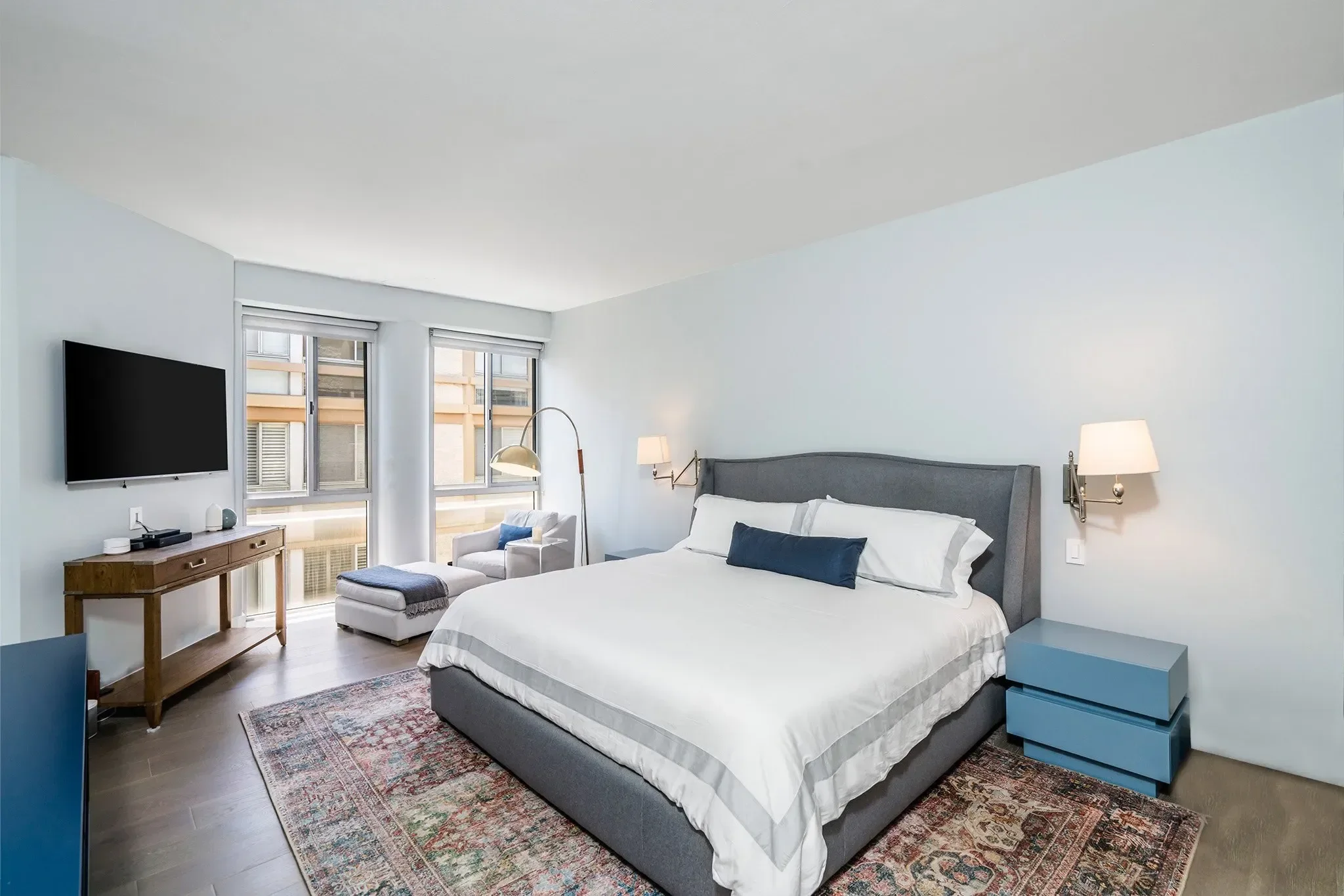A modern bedroom with a large bed, gray headboard, and white bedding with navy pillows. There are two nightstands with lamps, a wooden console table, and a white armchair near the window. The room has large windows with blinds, hardwood floors, and a