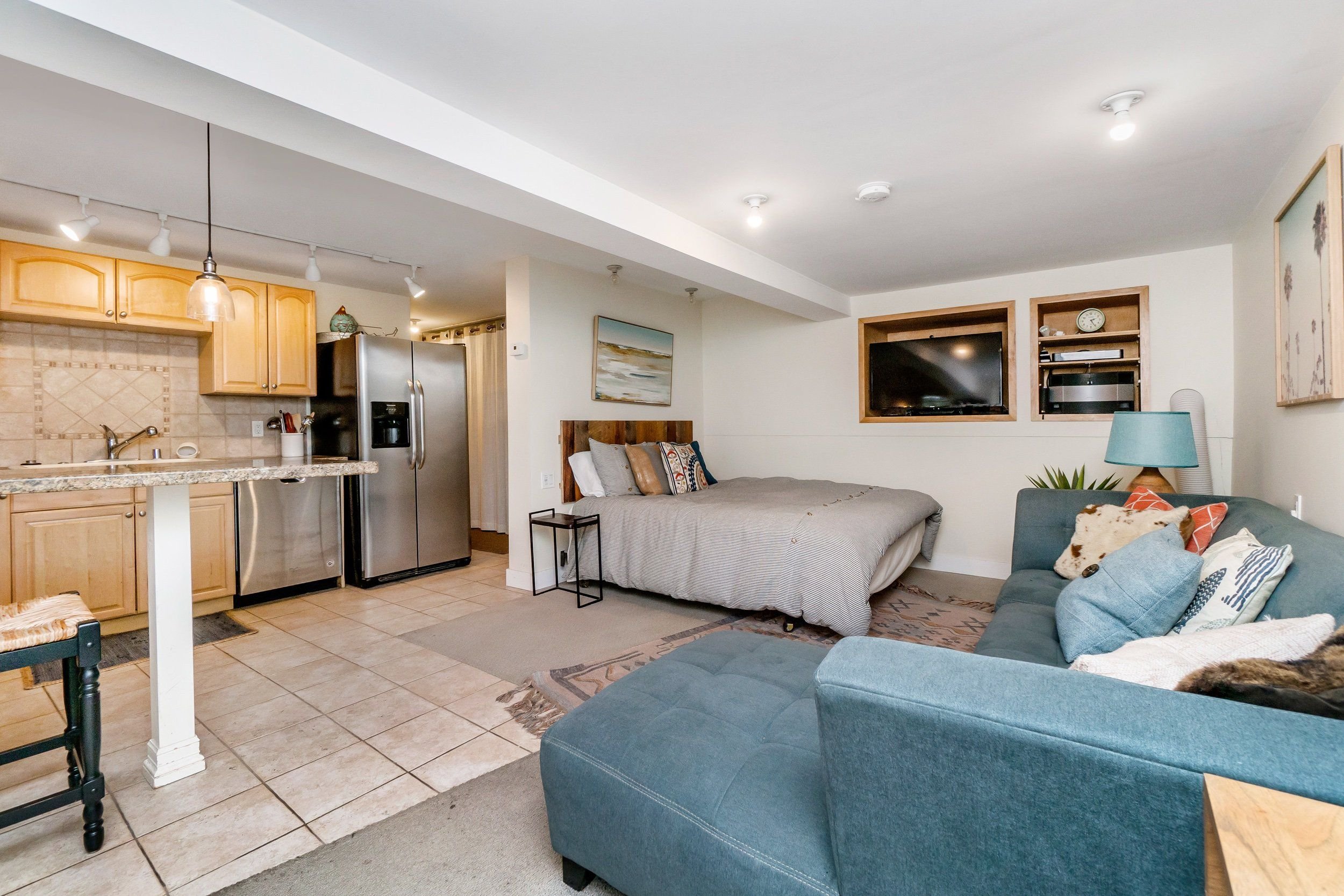Open-concept living area with a kitchen in the background. The kitchen has light wooden cabinets, a granite countertop, and stainless steel appliances, including a refrigerator. The living area features a bed with pillows, a blue sofa with decorative