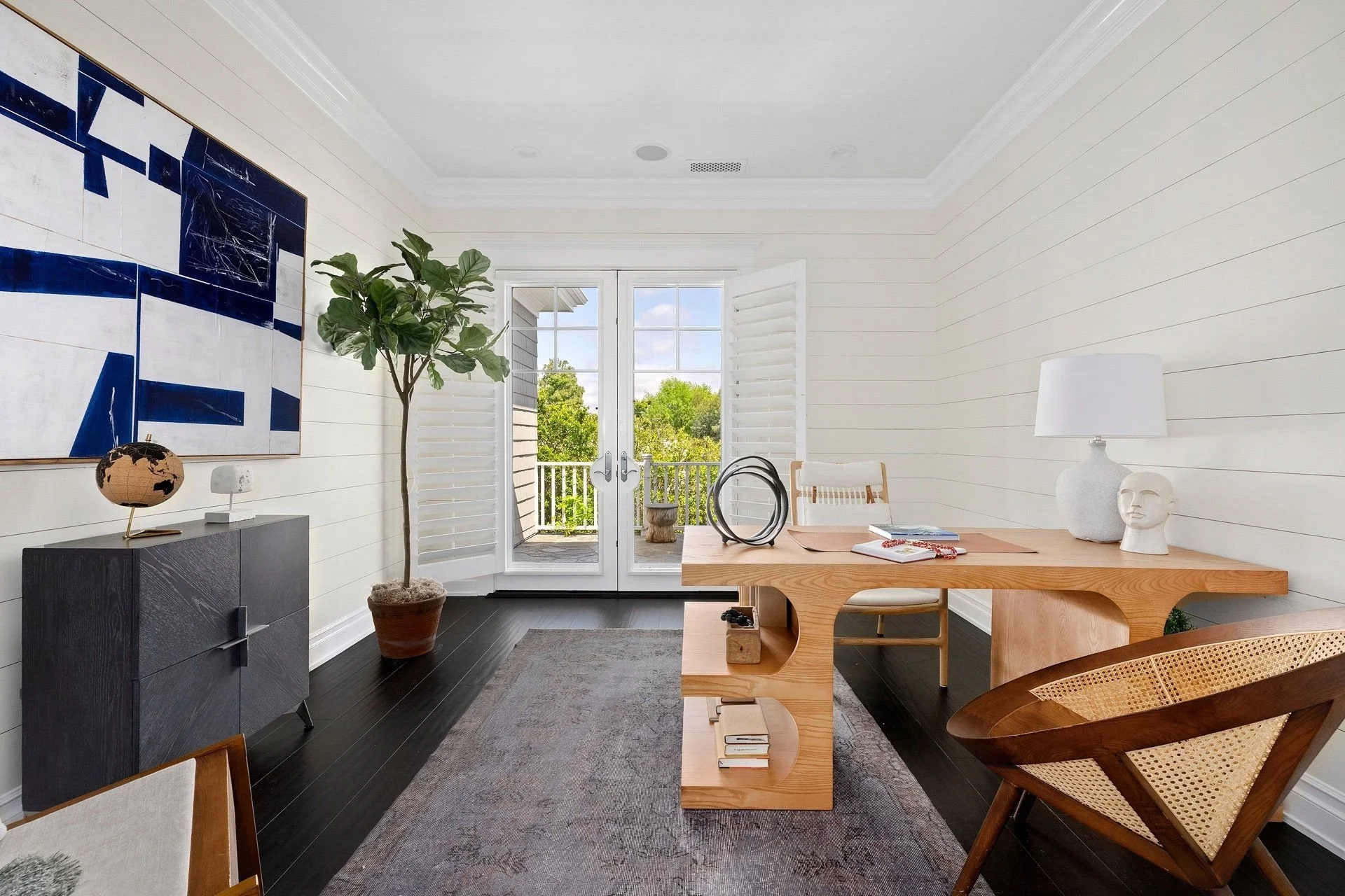 Bright living room with white shiplap walls, dark hardwood floors, a large windowed door leading to a balcony, a wooden dining table with modern decor, and a potted tree.