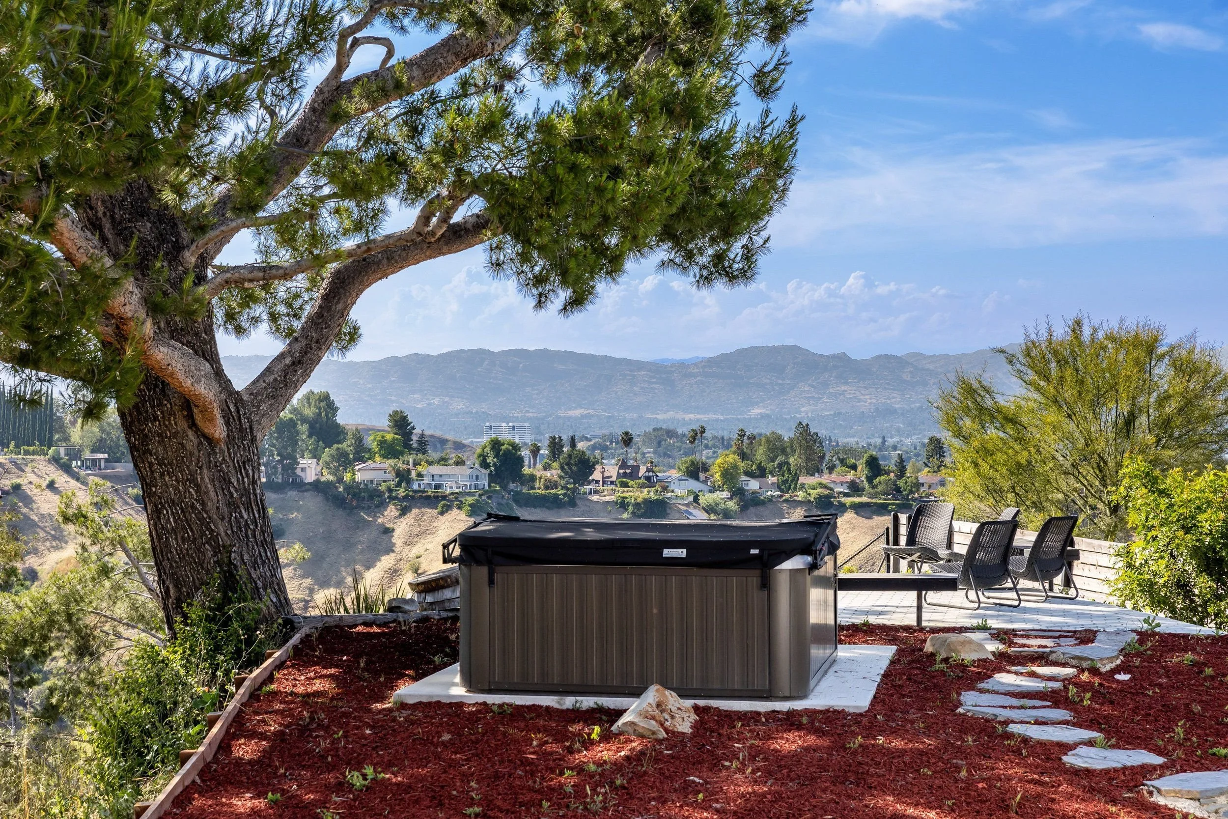 Large tree with green foliage on a hillside backyard with mountain views in the distance, outdoor chairs, and a storage shed on mulch-covered ground.