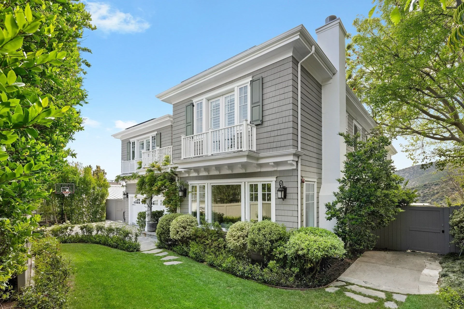 A two-story house with grey siding, white trim, and a small balcony on the second floor. The front yard is lush with green grass, shrubs, and trees, and there is a basketball hoop on the left side of the yard. The sky is blue with some clouds.