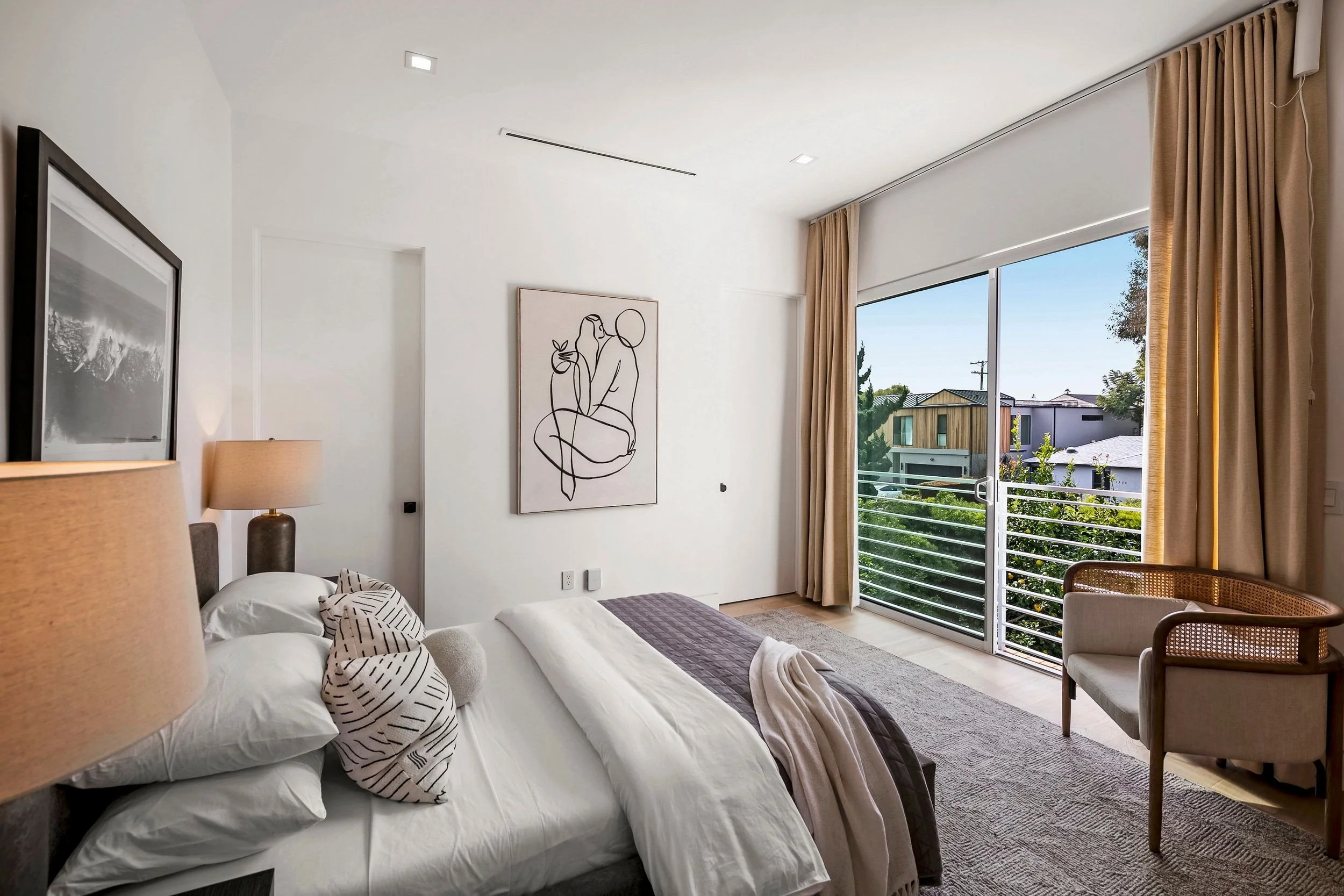 A modern bedroom with a large bed decorated with black and white blankets and pillows. The room features a large sliding glass door opening to a balcony with a view of neighboring houses and trees. The walls are white with minimalistic art and two ta