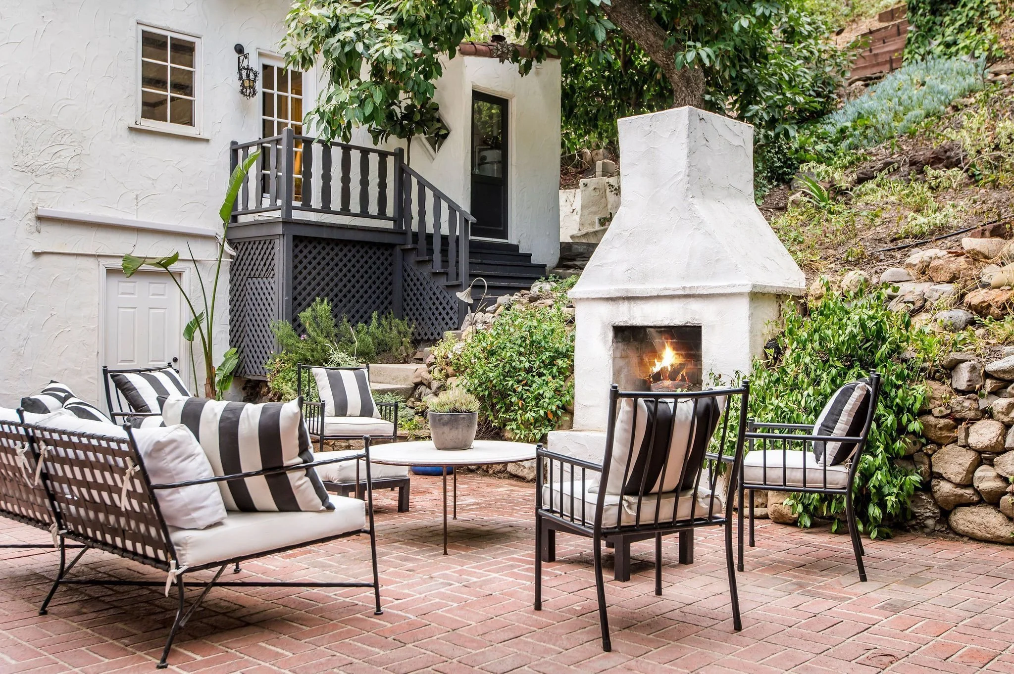 An outdoor patio with a brick floor, black metal chairs with striped cushions, a white potted plant, a white fireplace with a fire burning, and a white stucco house with a small deck and three windows surrounded by green foliage.