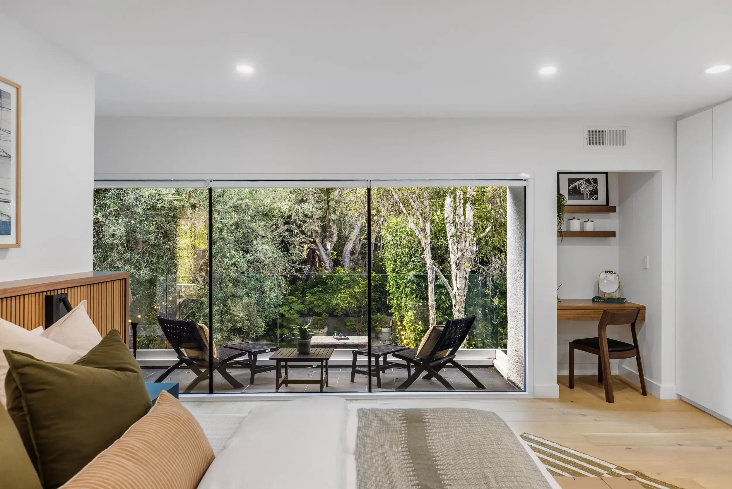 Living room with a large window overlooking a backyard with trees, outdoor chairs, and small patio table, decorated with indoor plants and framed black and white art on the wall.