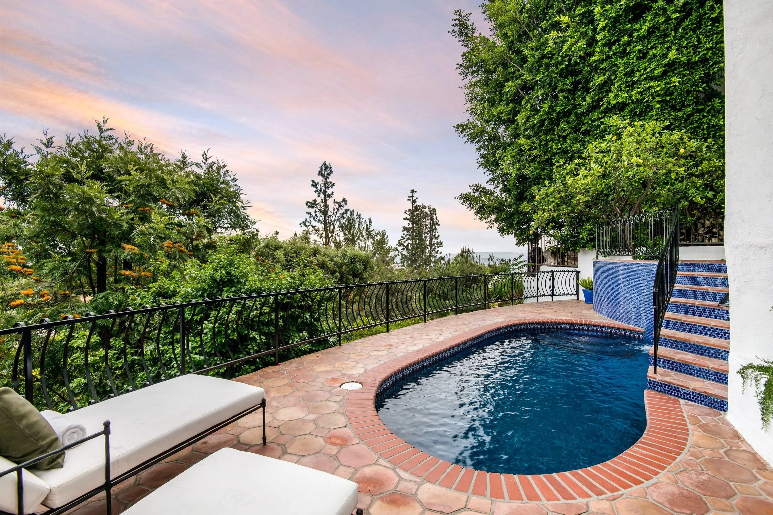 A backyard swimming pool with a curved edge, tile interior, and brick coping. There are white lounge chairs with rolled towels on a stone-tiled patio. The area is enclosed by a black metal fence, surrounded by lush green trees and plants. The sky abo