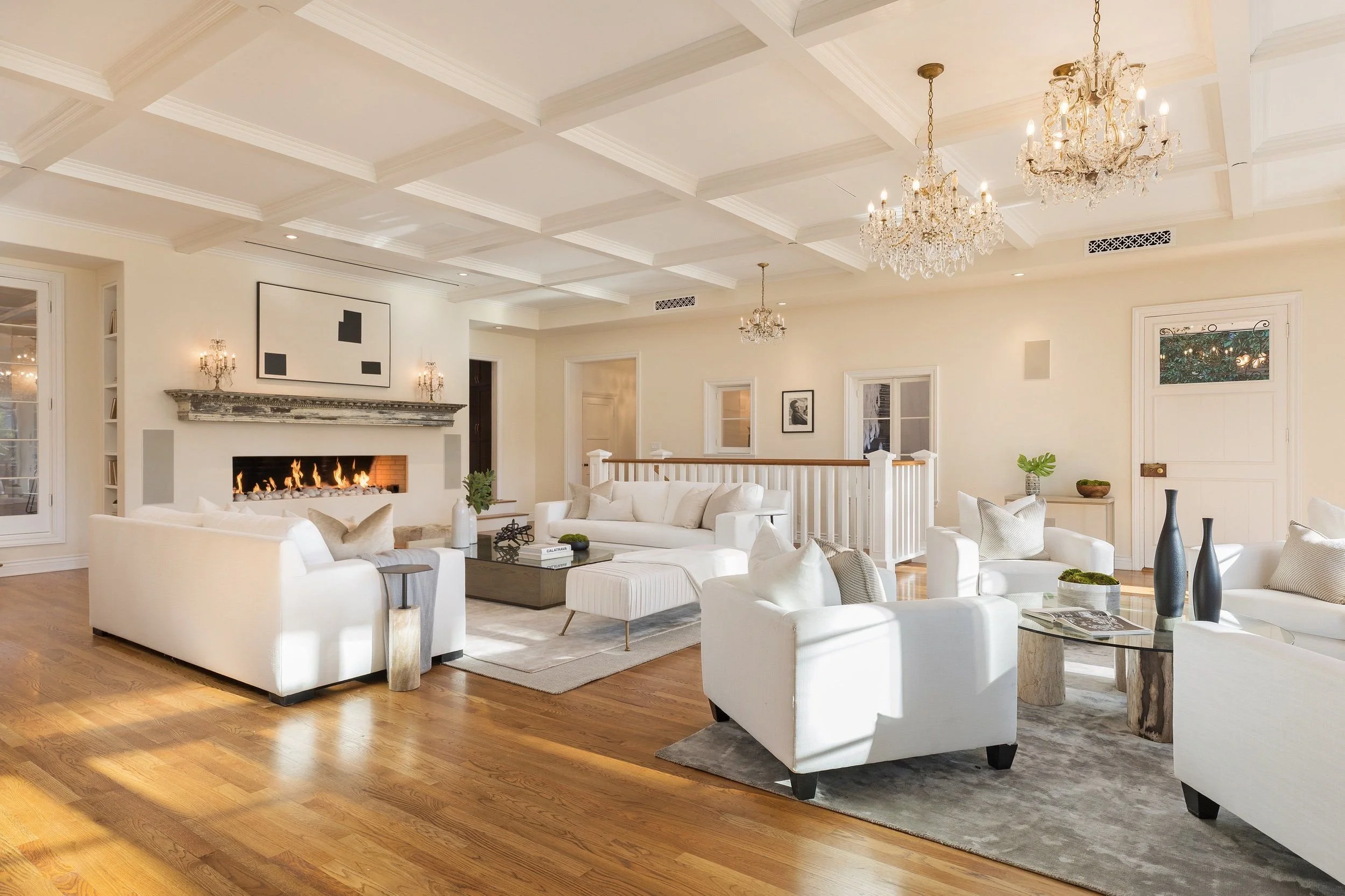 A spacious living room with white sofas, a fireplace, and a coffered ceiling with chandeliers, hardwood flooring, and decorative wall art and plants.