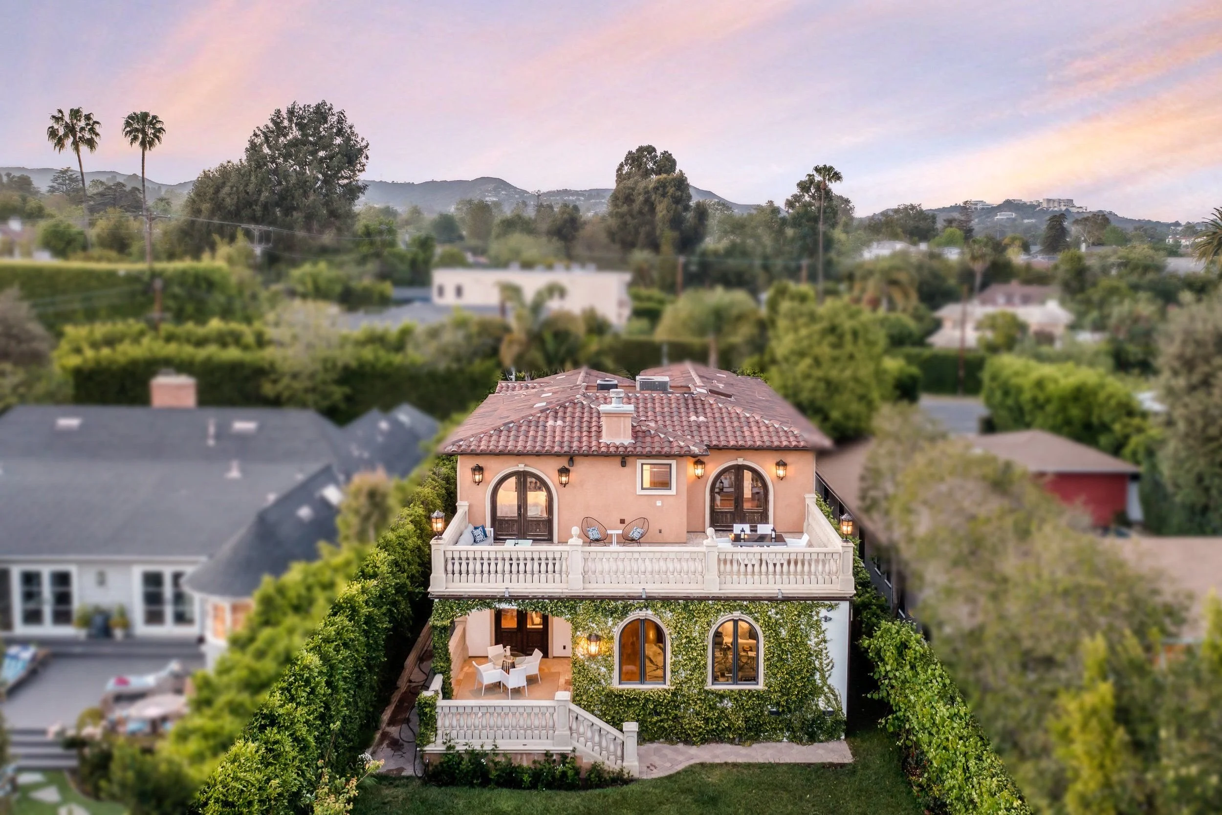A multi-story house with a pink exterior and balcony, surrounded by greenery, with a mountain range in the background during sunset.
