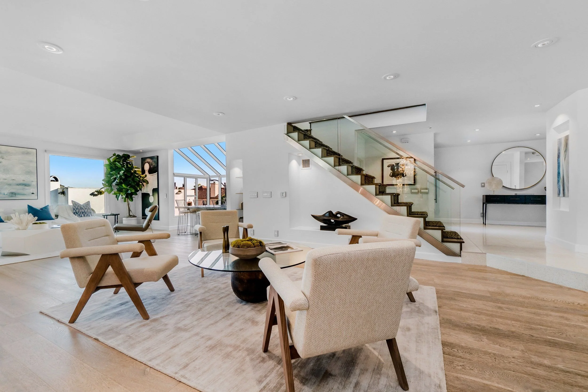 Spacious, modern living room with white walls and ceiling, wooden flooring, beige and white furniture, a glass staircase, large windows, and decor including artwork, plants, and mirrors.