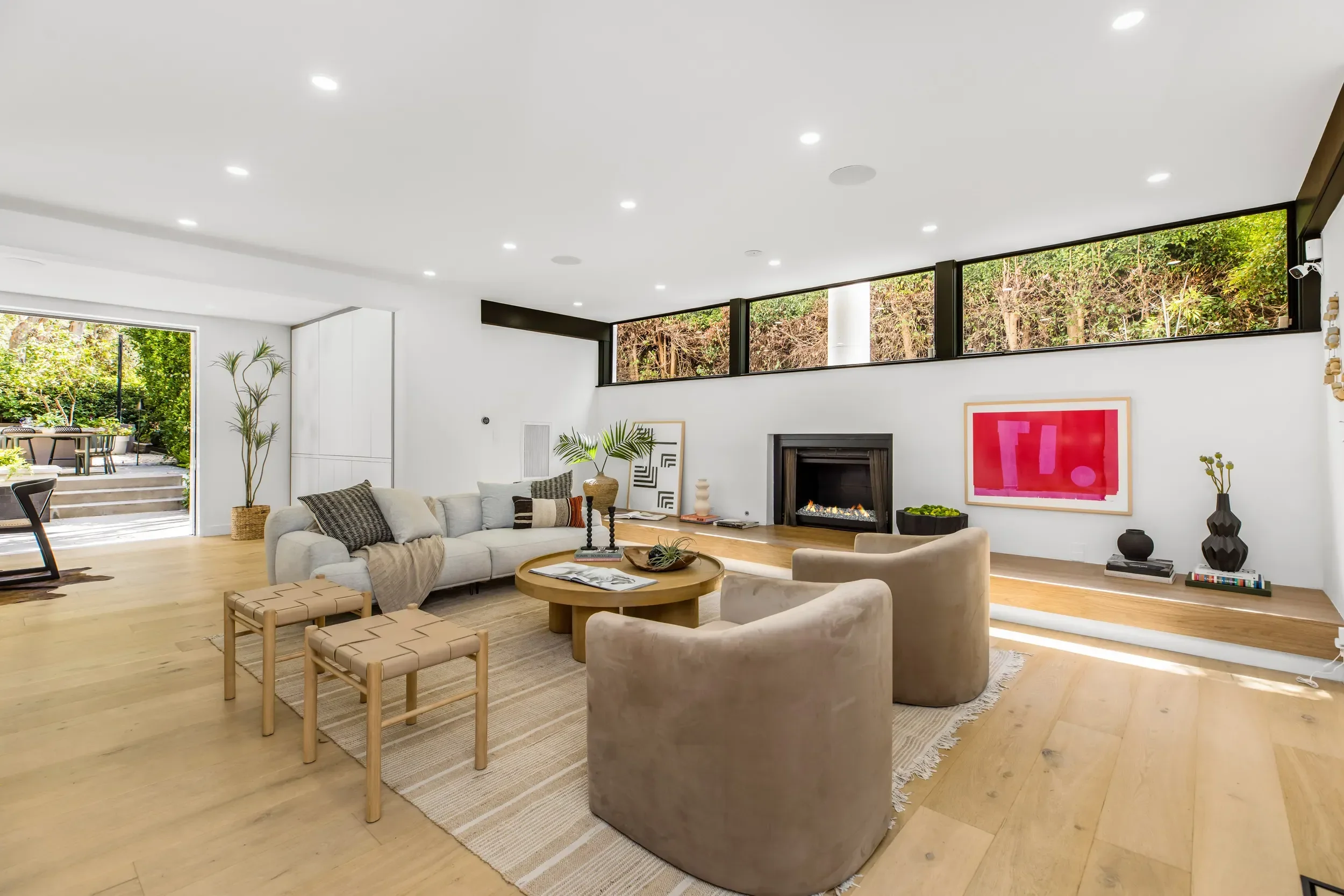 Modern living room with a white ceiling and walls, large windows, and light wooden floors. Contains white sofas, a round wooden coffee table, and decorative plants. There's a fireplace with a pink framed artwork above it, and outside are green trees 