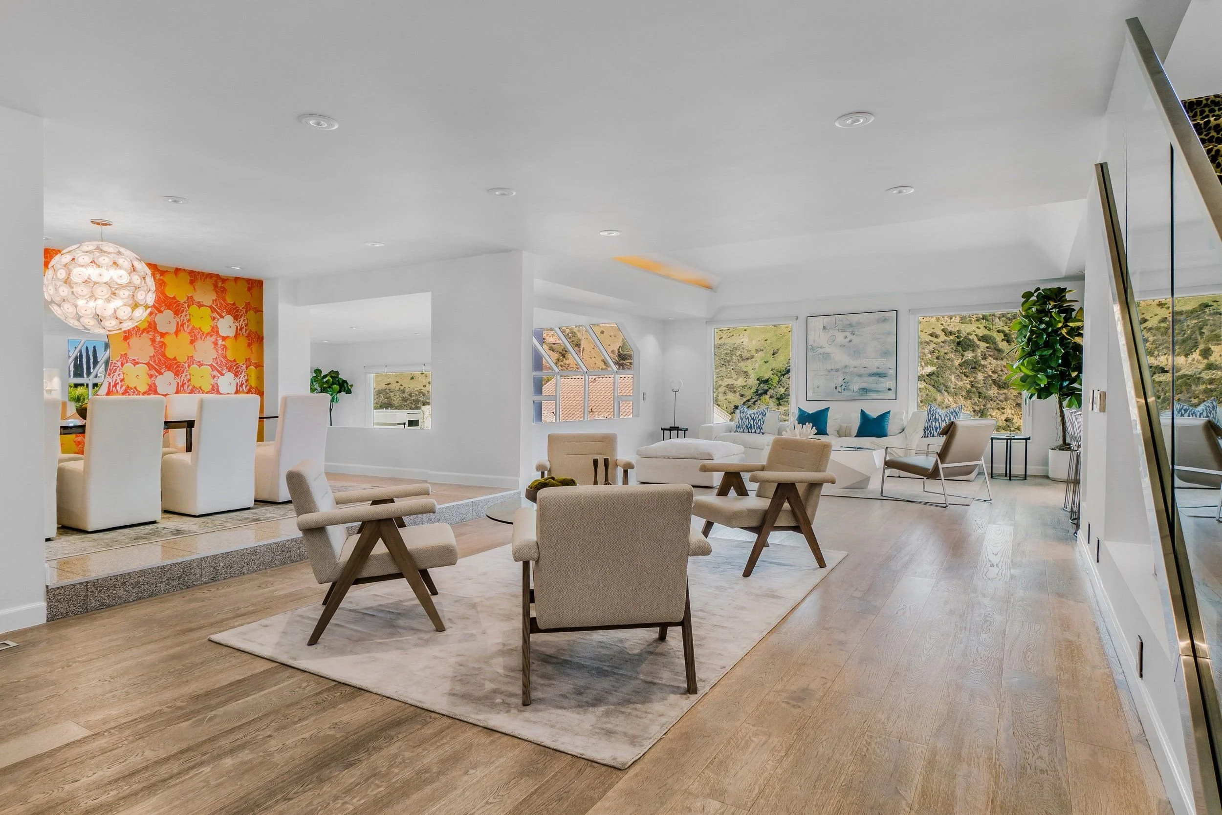 Interior of a modern, bright living room with white walls, large windows, wooden flooring, beige and white furniture, decorative plants, and artwork on the walls.