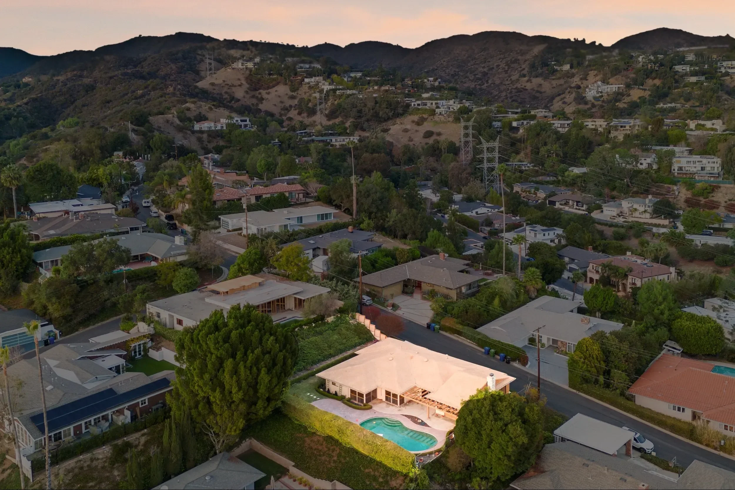 An aerial view of a residential neighborhood with houses, trees, and a hillside in the background. There is a swimming pool in the backyard of one house.