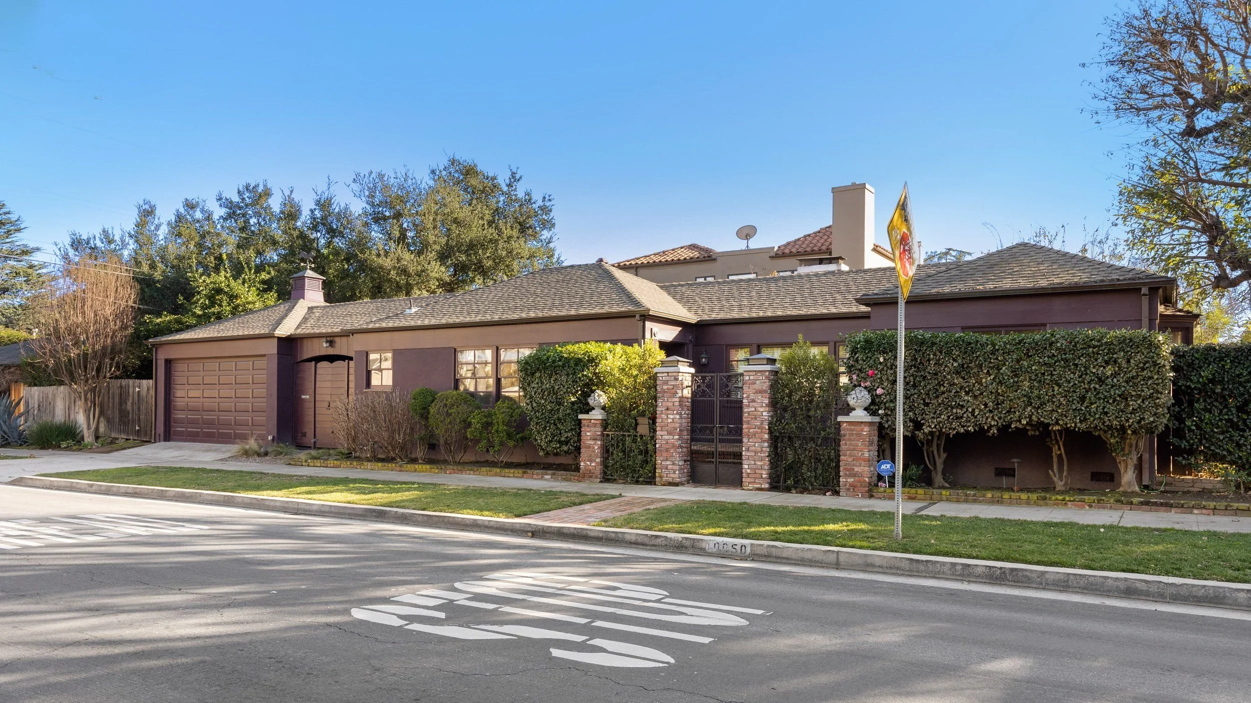 Residential house with a brown exterior, a garage door, brick and iron gate, landscaped front yard with bushes and trees, street view with crosswalk and sidewalk, clear blue sky.