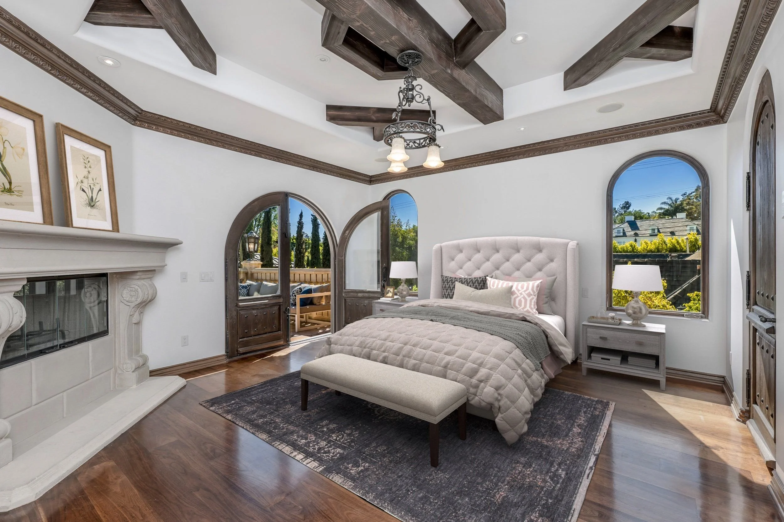 A bedroom with white walls, hardwood floors, and dark wood ceiling beams. It features a queen-sized bed with an upholstered headboard, patterned pillows, and a quilted blanket. There are nightstands with lamps on each side of the bed and a bench at t