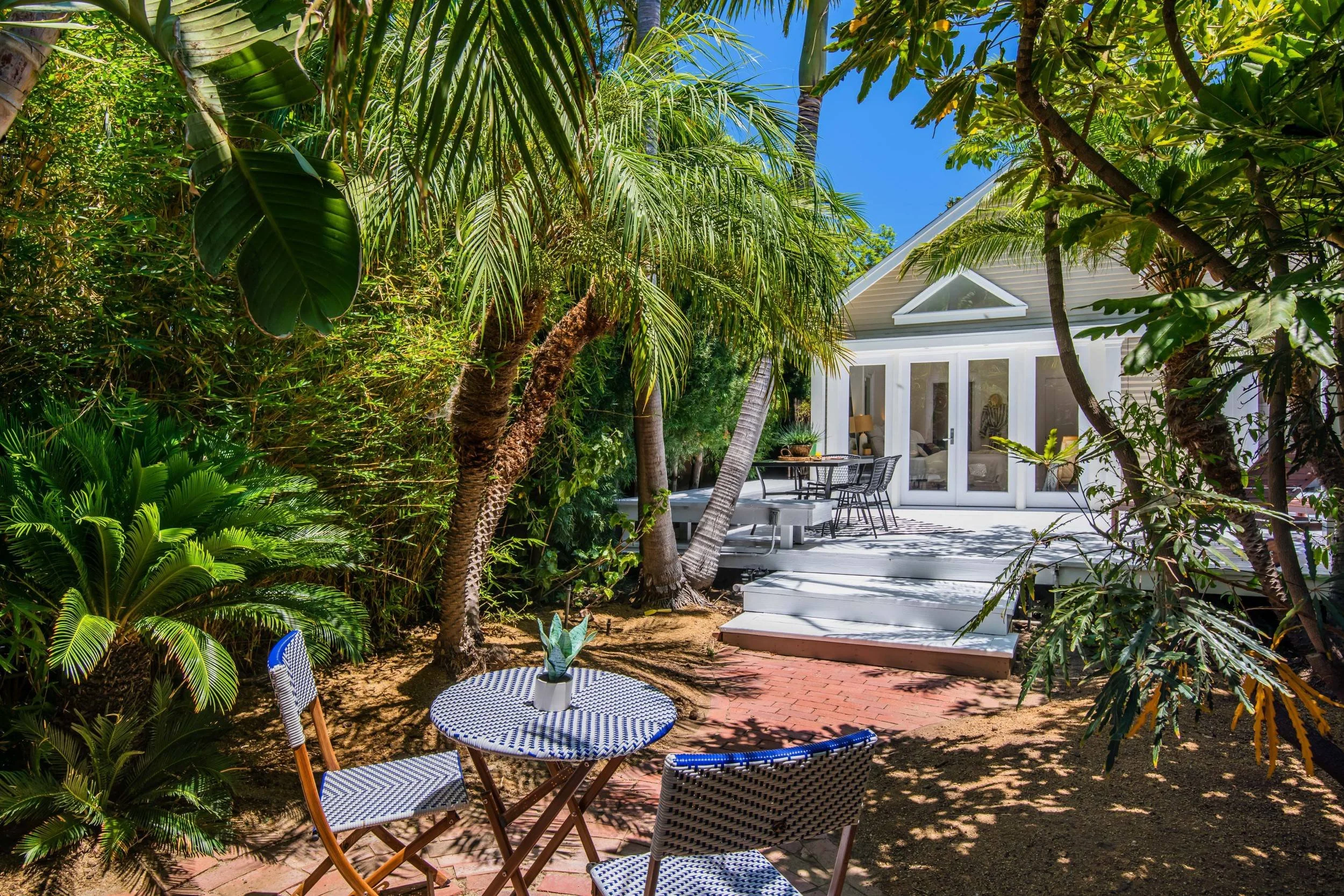 A backyard with lush tropical plants and trees surrounding a white house with a covered porch. The patio area has outdoor furniture, including a round table with a potted plant and several chairs. The scene is sunny with a bright blue sky overhead.