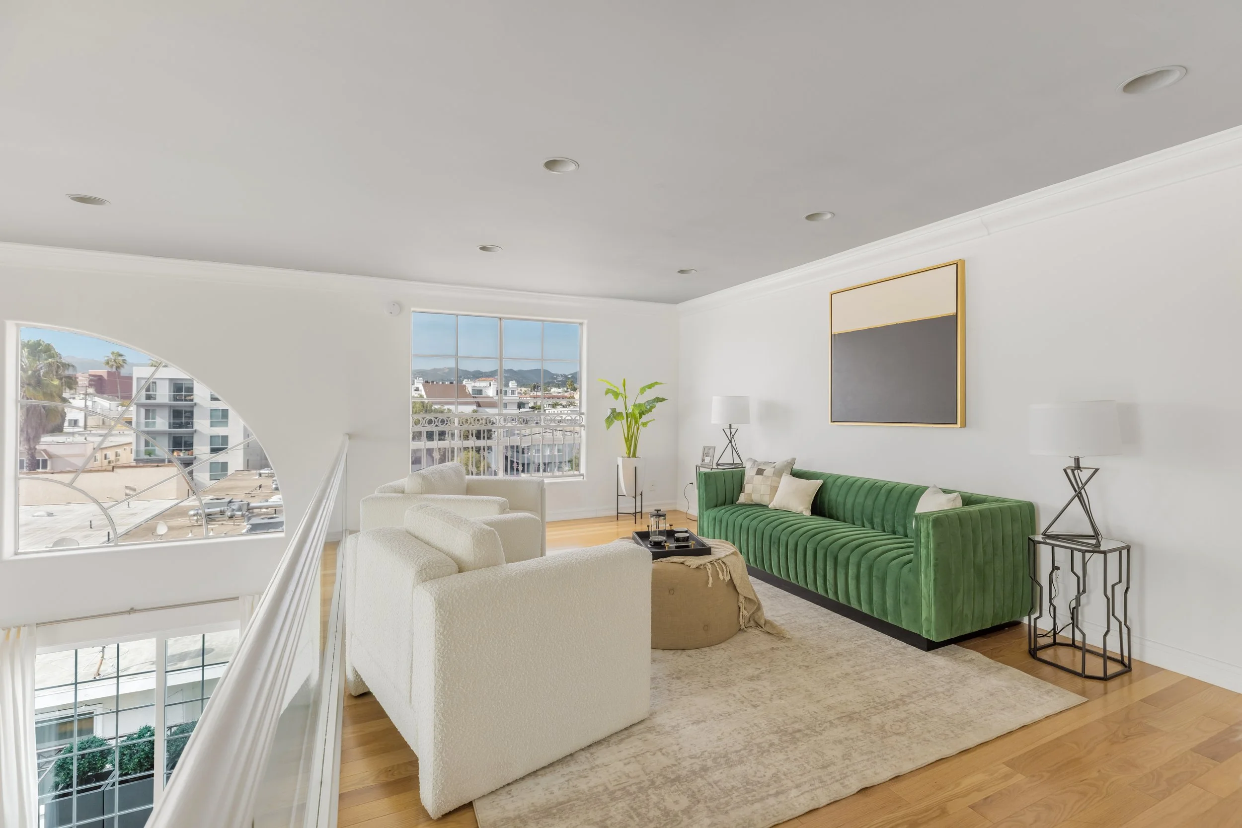 Living room with large window, white walls, and a green velvet sofa. There are two white armchairs, a beige ottoman, a black tray with candles, and two tables with lamps. Artwork hangs on the wall, and a view of neighboring buildings and mountains is