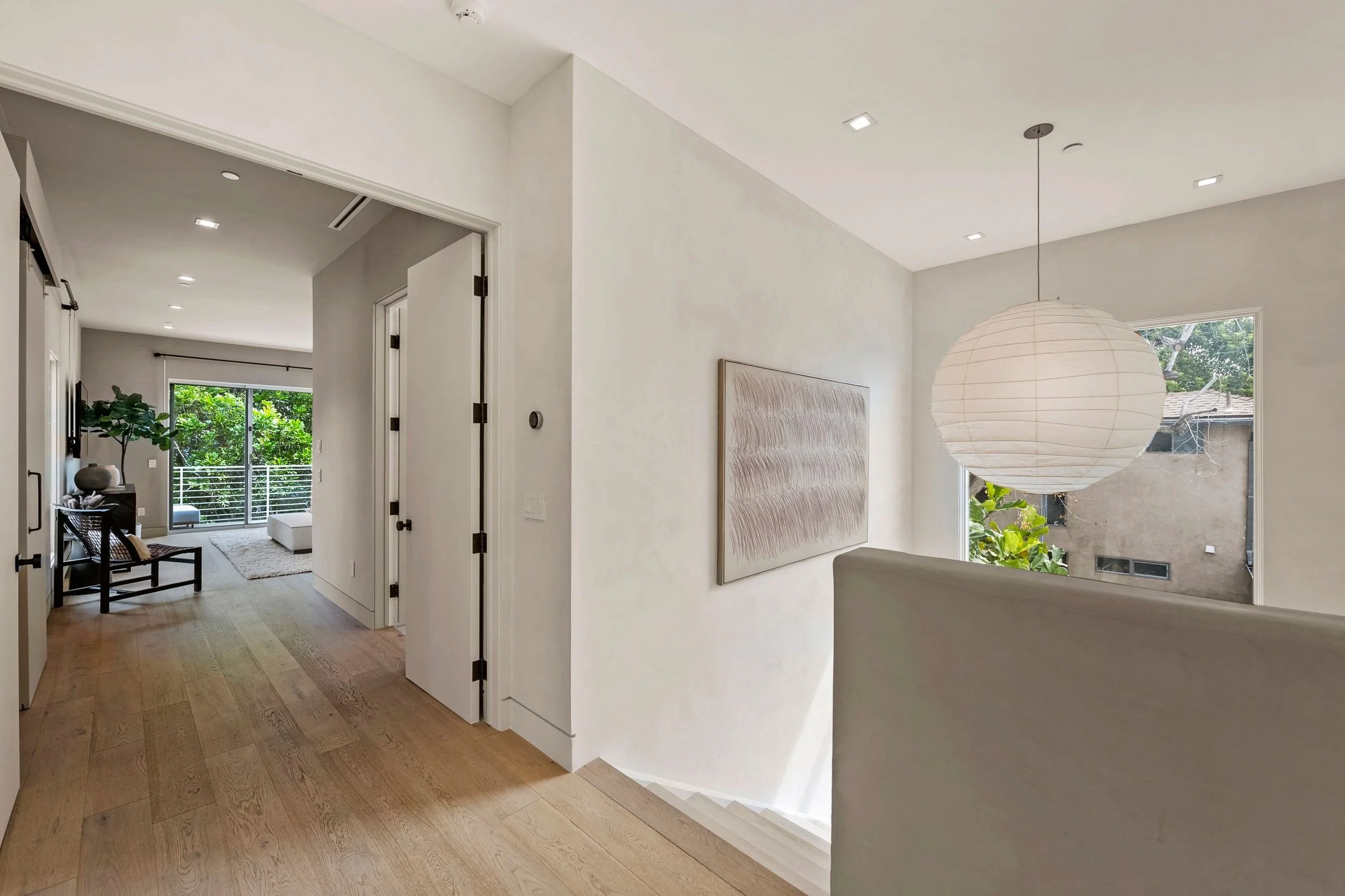 A bright hallway leading to a living room with a sliding glass door overlooking green plants outside. The hallway has light wooden flooring, white walls, and an open doorway with black hinges. There's a simple black chair with cushions on it near the