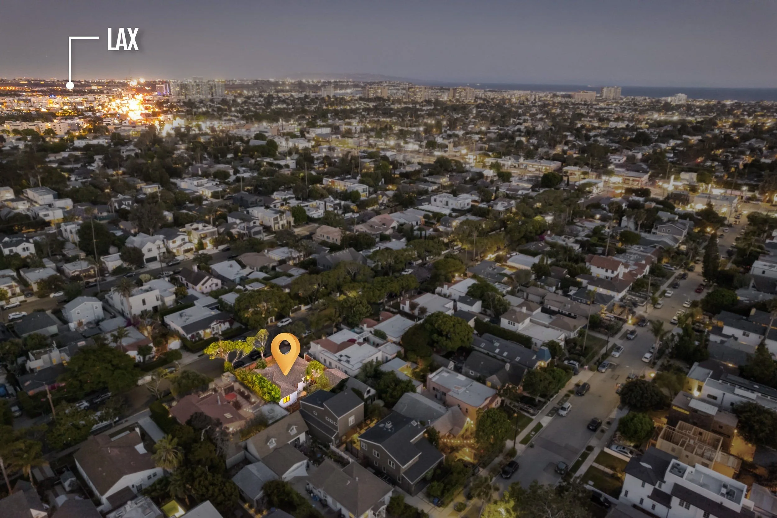 An aerial nighttime view of a city with many houses, trees, and streets illuminated by streetlights. The background shows taller buildings and a coastline. There is a location marker icon on one of the houses in the foreground.
