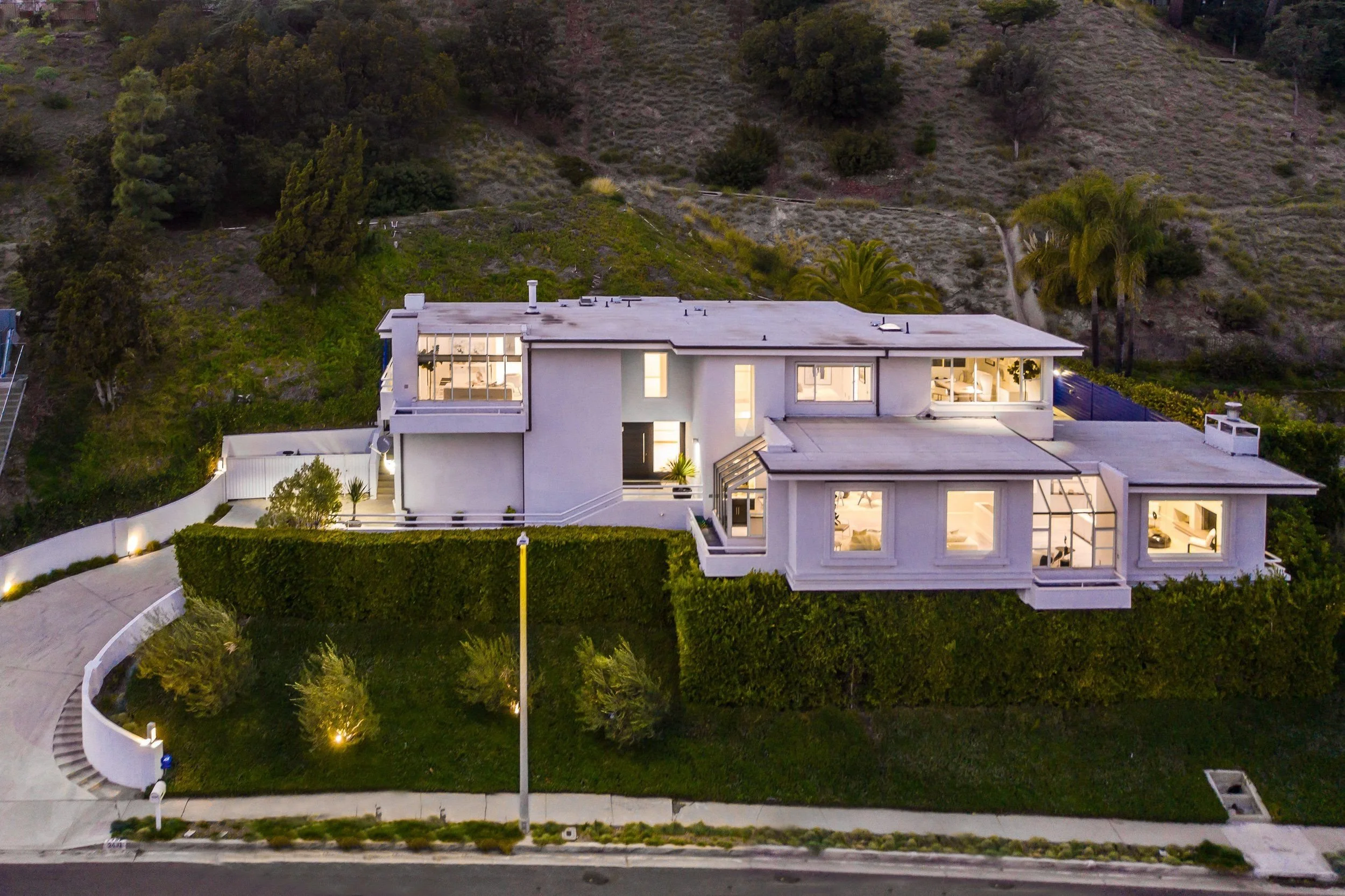 A modern house with large windows and a flat roof, illuminated from inside, surrounded by greenery and a curved white fence, with a hillside covered in bushes and trees in the background during dusk.