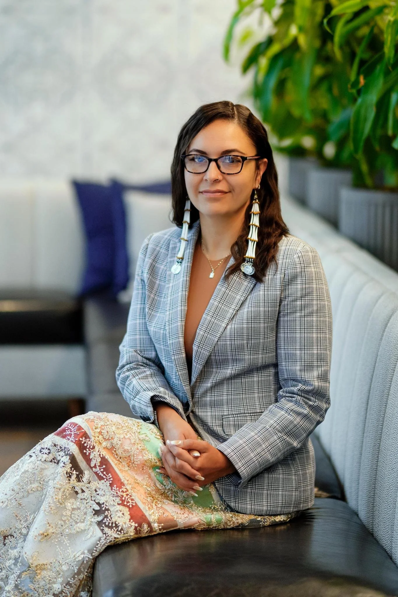 A woman with long dark hair, glasses, and large earrings sitting on a bench with plants behind her, wearing a plaid blazer and a colorful patterned skirt.