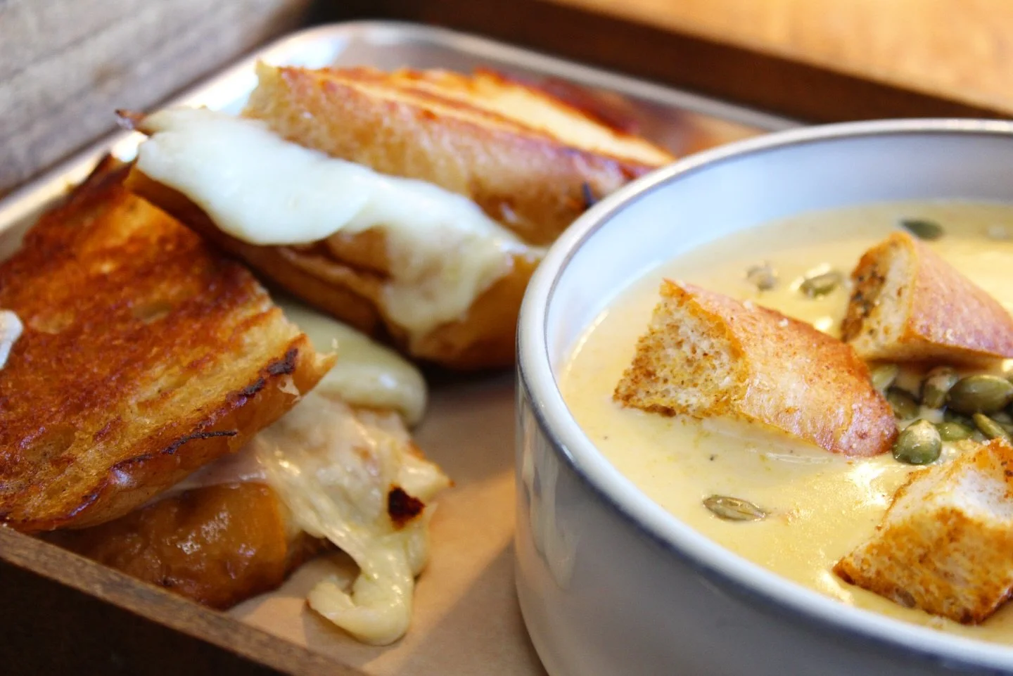 Close-up of a lunch tray with grilled cheese sandwiches, one with melted cheese and the other with melted cheese and a fried egg, and a bowl of creamy soup with croutons and pumpkin seeds.