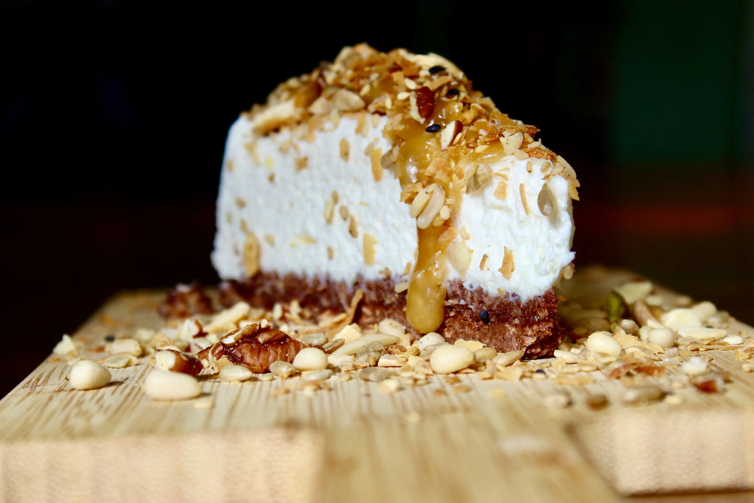 A slice of cheesecake topped with chopped nuts and caramel, placed on a wooden surface surrounded by nuts and crumbs.